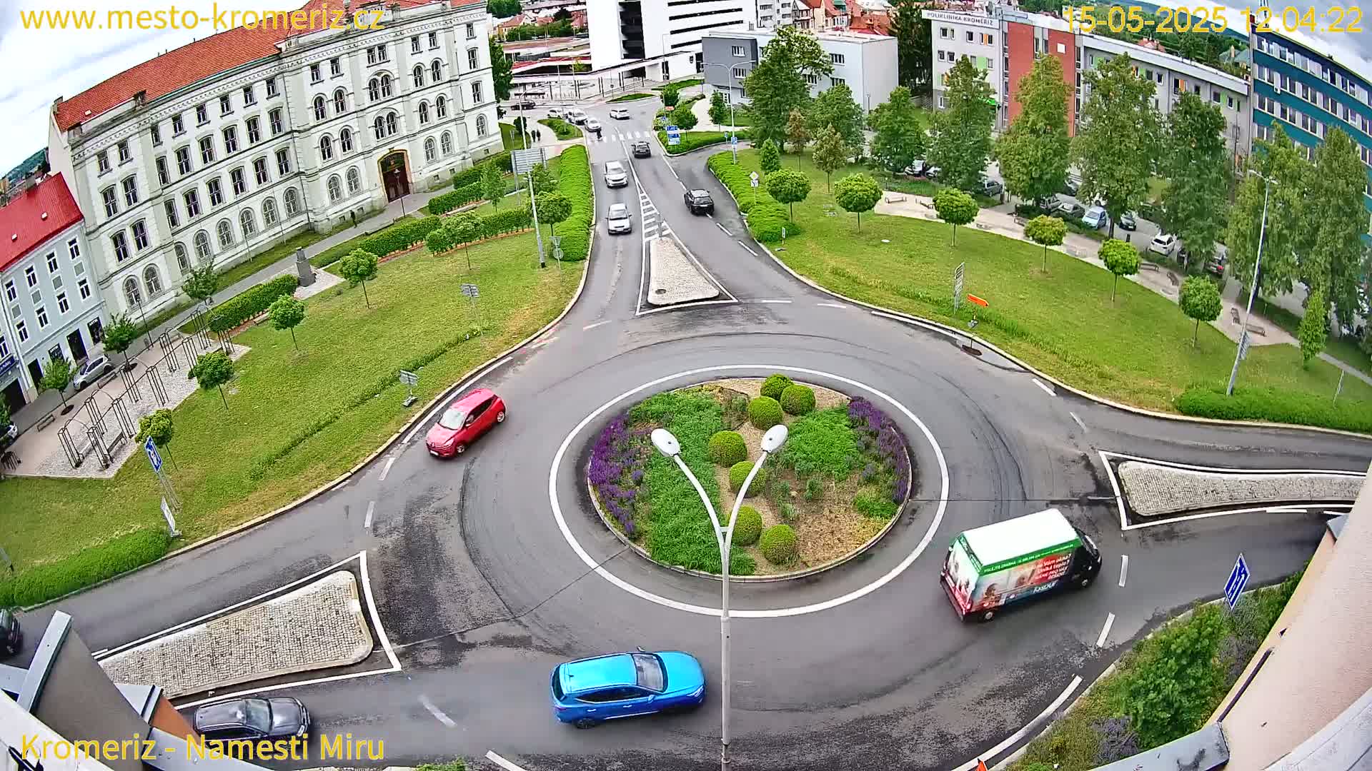 A roundabout surrounded by a grassy area with buildings and several cars, including a red car, a blue car, and a white van, on a partly cloudy day.