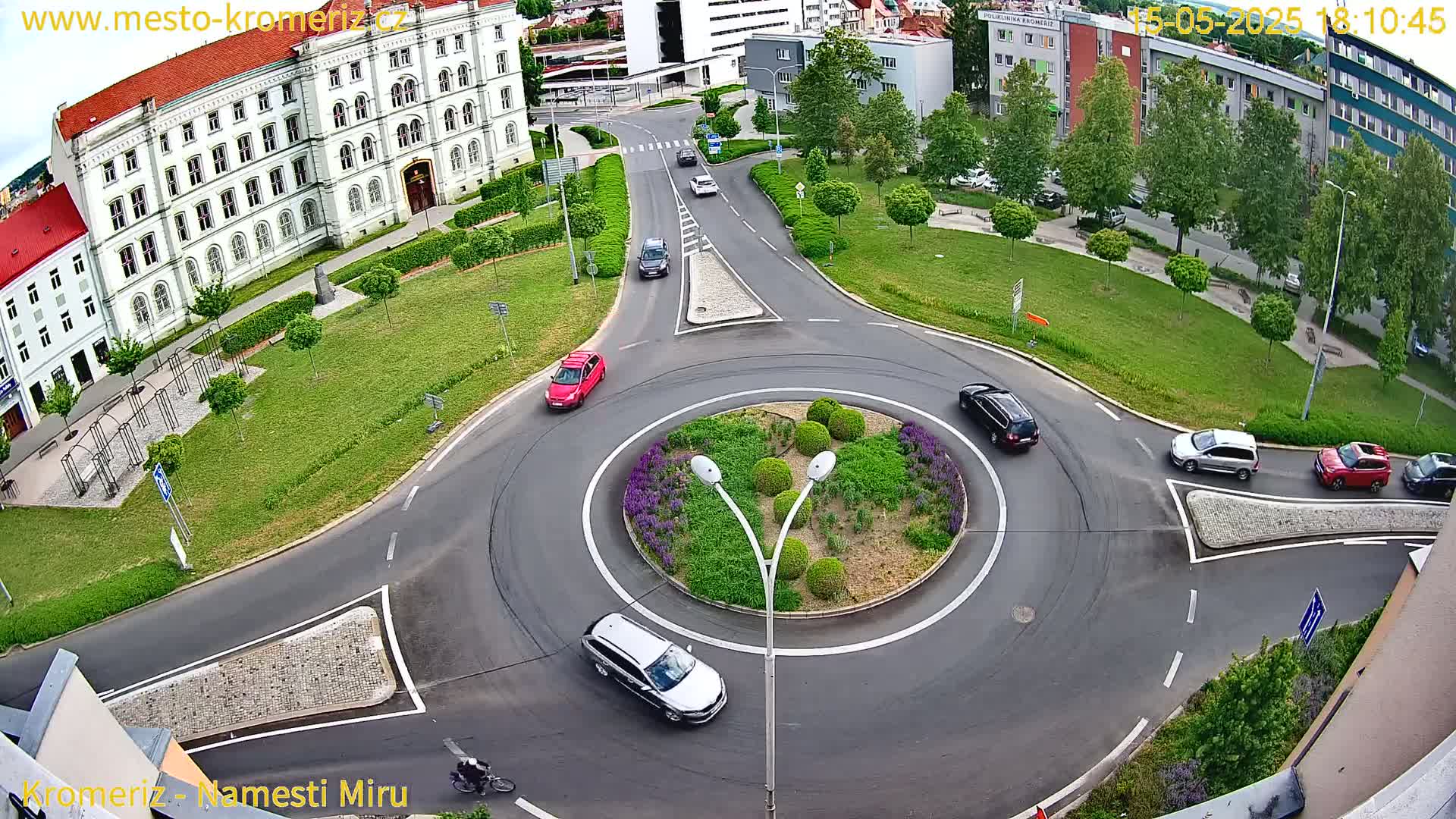 A high-angle view of a roundabout with several cars navigating it, surrounded by buildings and green spaces on a partly sunny day.