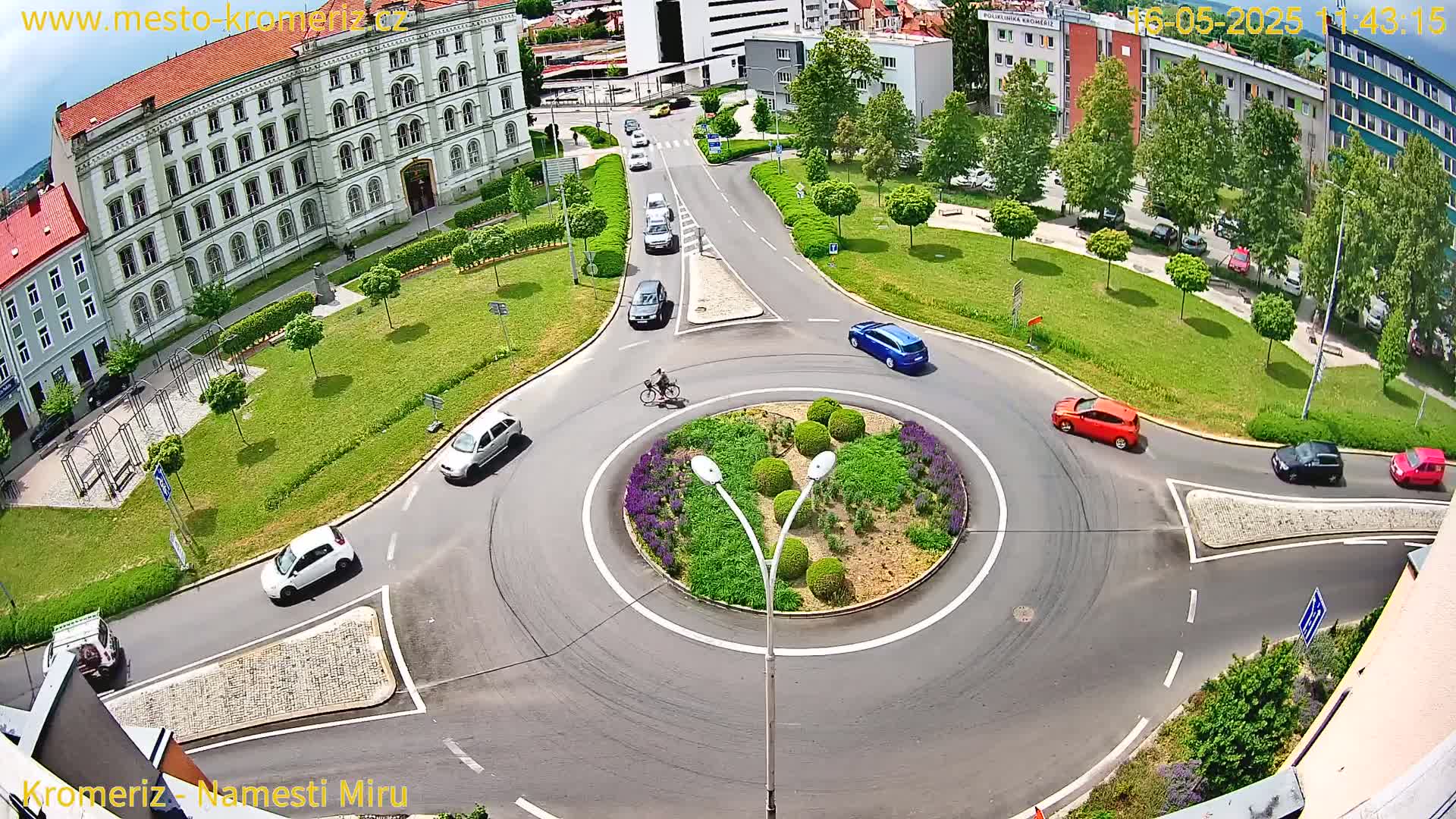 A sunny aerial view of a roundabout surrounded by a landscaped area with buildings and cars driving on roads.