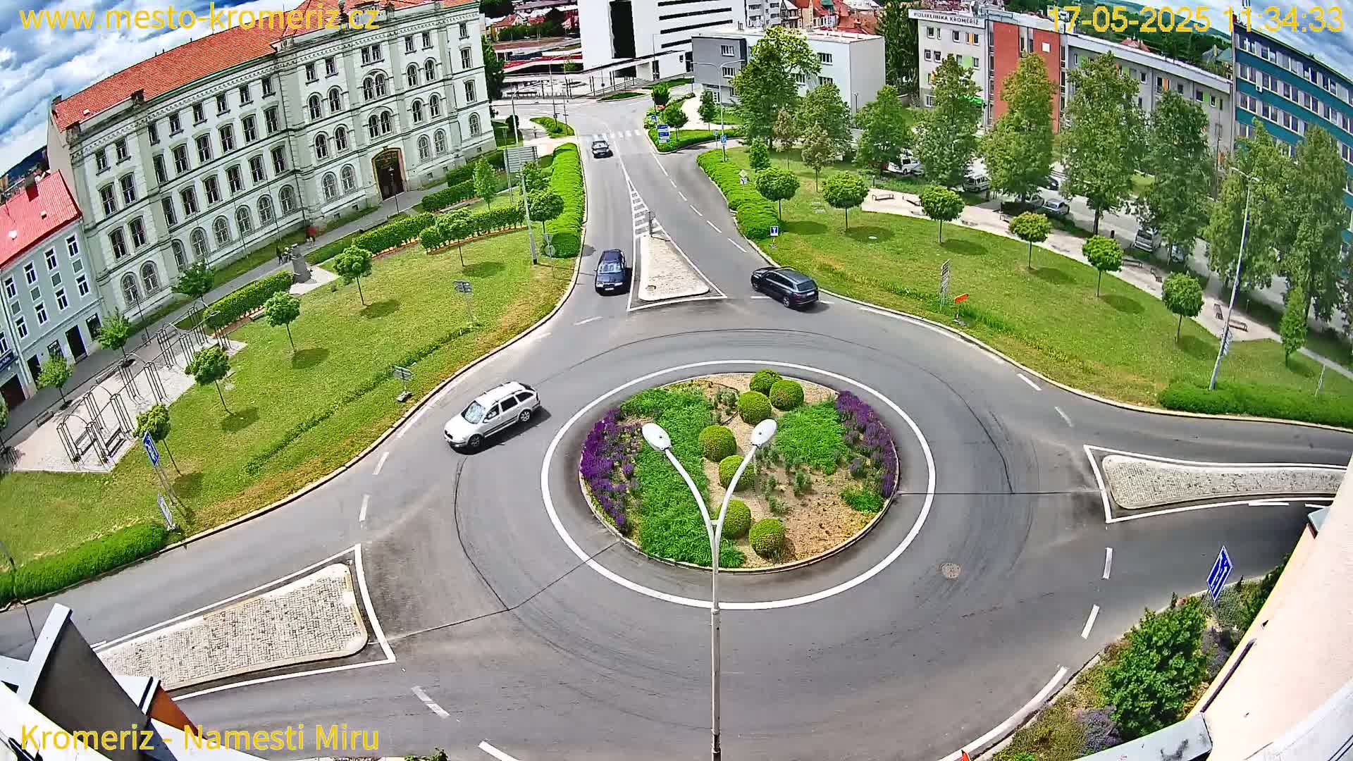 A high-angle view of a roundabout surrounded by green lawns and buildings on a sunny day, with several cars navigating the traffic circle.