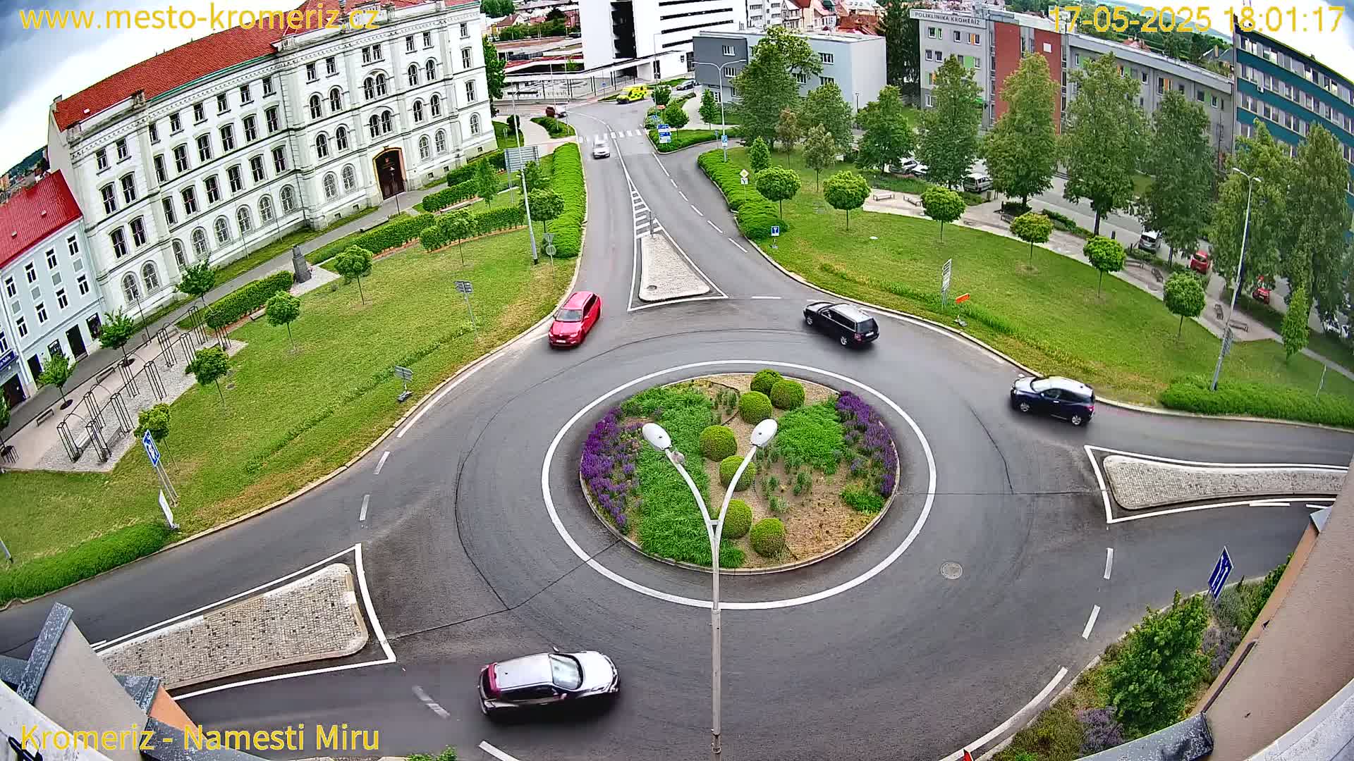 An aerial view shows a roundabout with several cars driving around a landscaped center island, surrounded by a large building, grassy areas, and trees under a clear sky.
