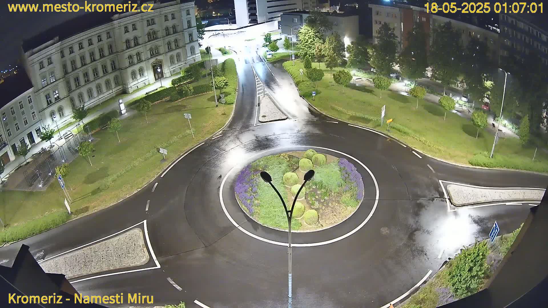 A wet roundabout at night, surrounded by a large building and green spaces, is illuminated by streetlights.