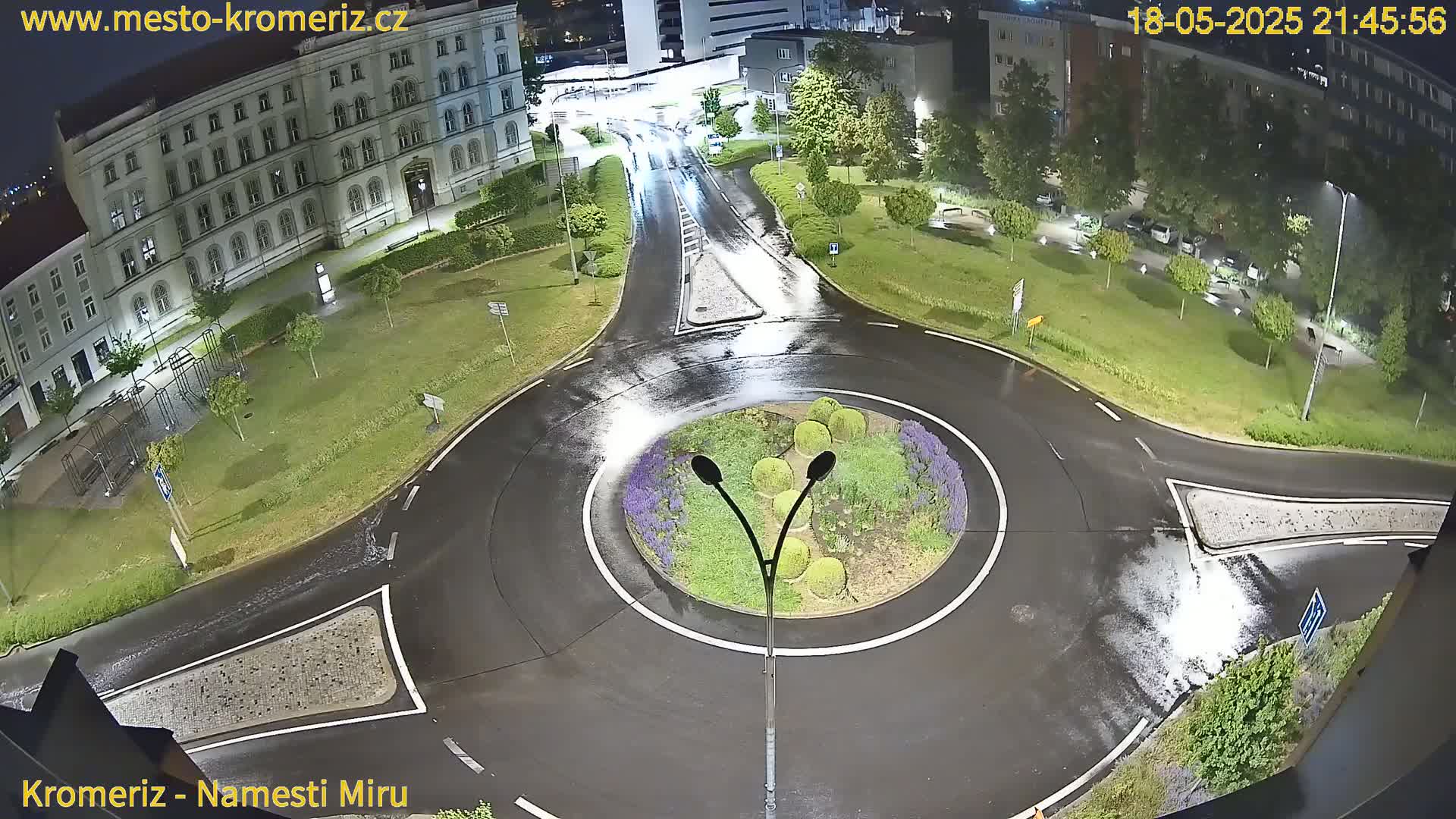 A wet roundabout and surrounding streets at night, with a large building and greenery visible.