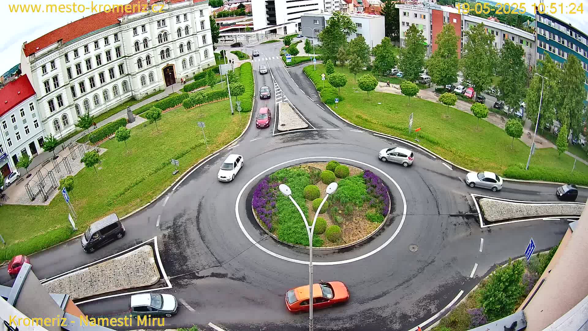 A wet roundabout surrounded by a grassy area, buildings, and several cars driving on a rainy day.