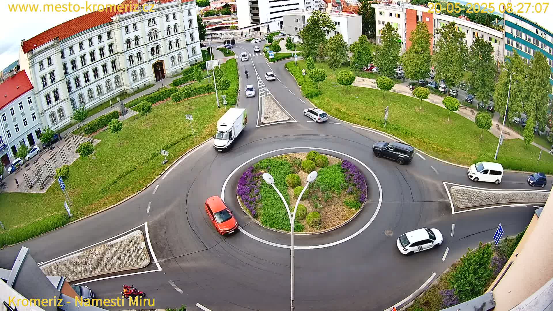 An aerial view of a roundabout with several cars and a delivery truck, surrounded by buildings and green spaces on a partly sunny day.