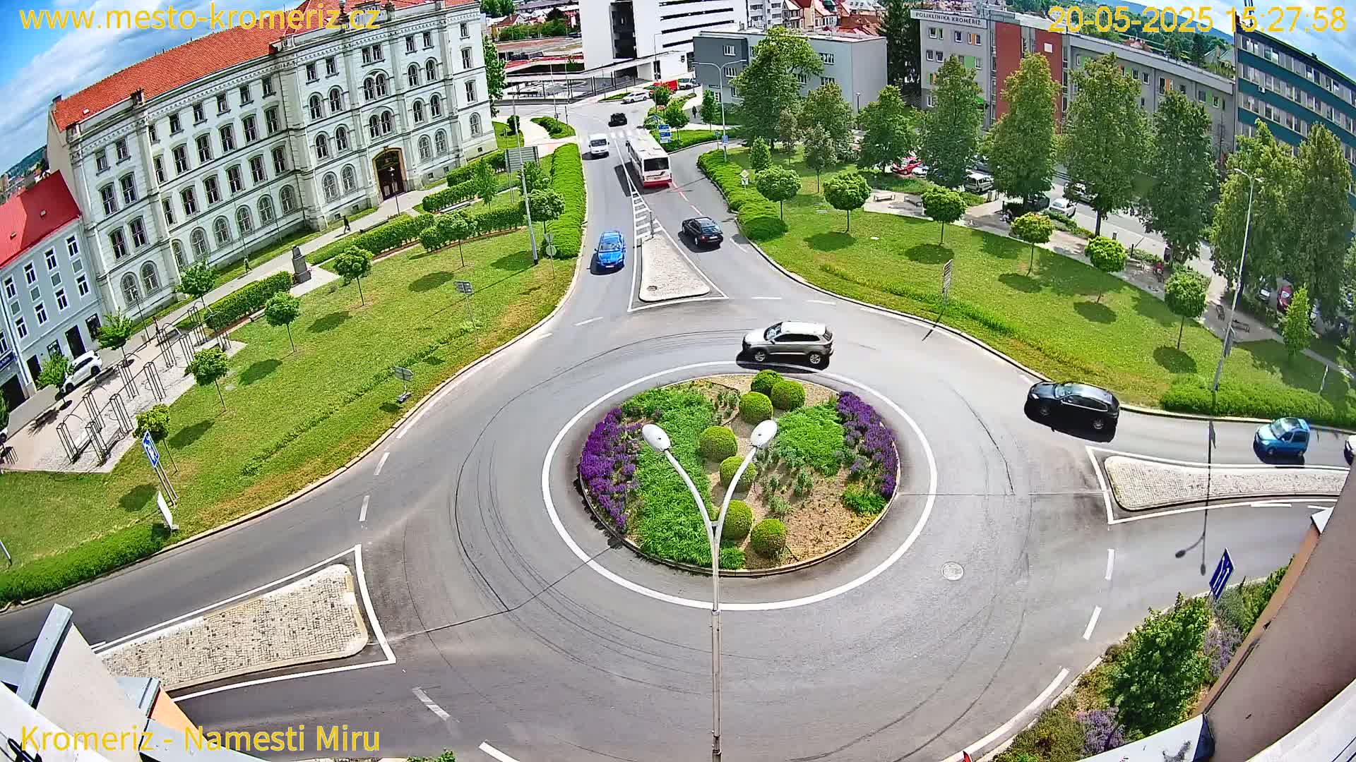 A sunny day shows a traffic circle surrounded by a grassy area, buildings, and several cars.