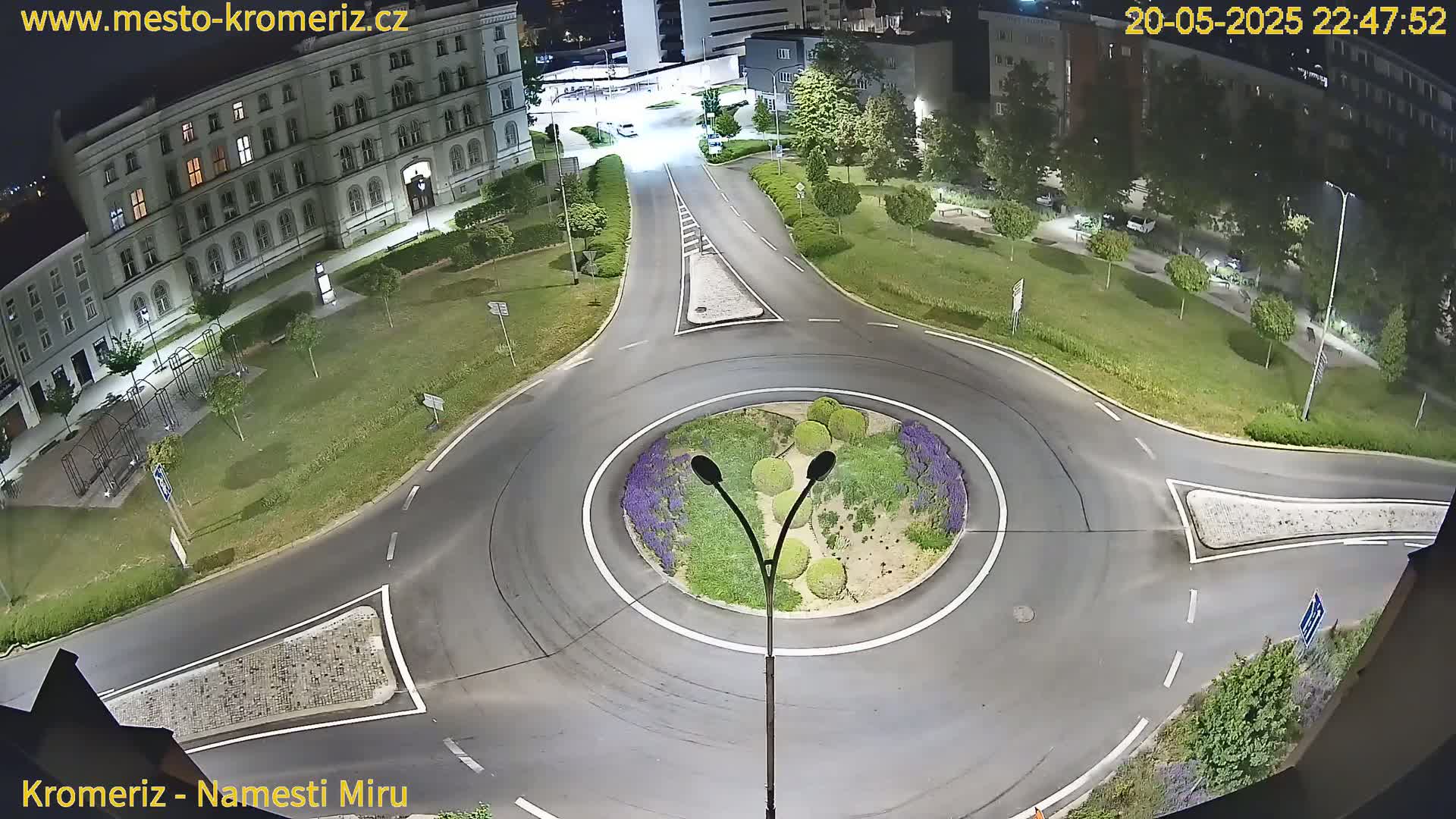 An elevated view at night shows a traffic circle surrounded by a grassy area, buildings, and streetlights.