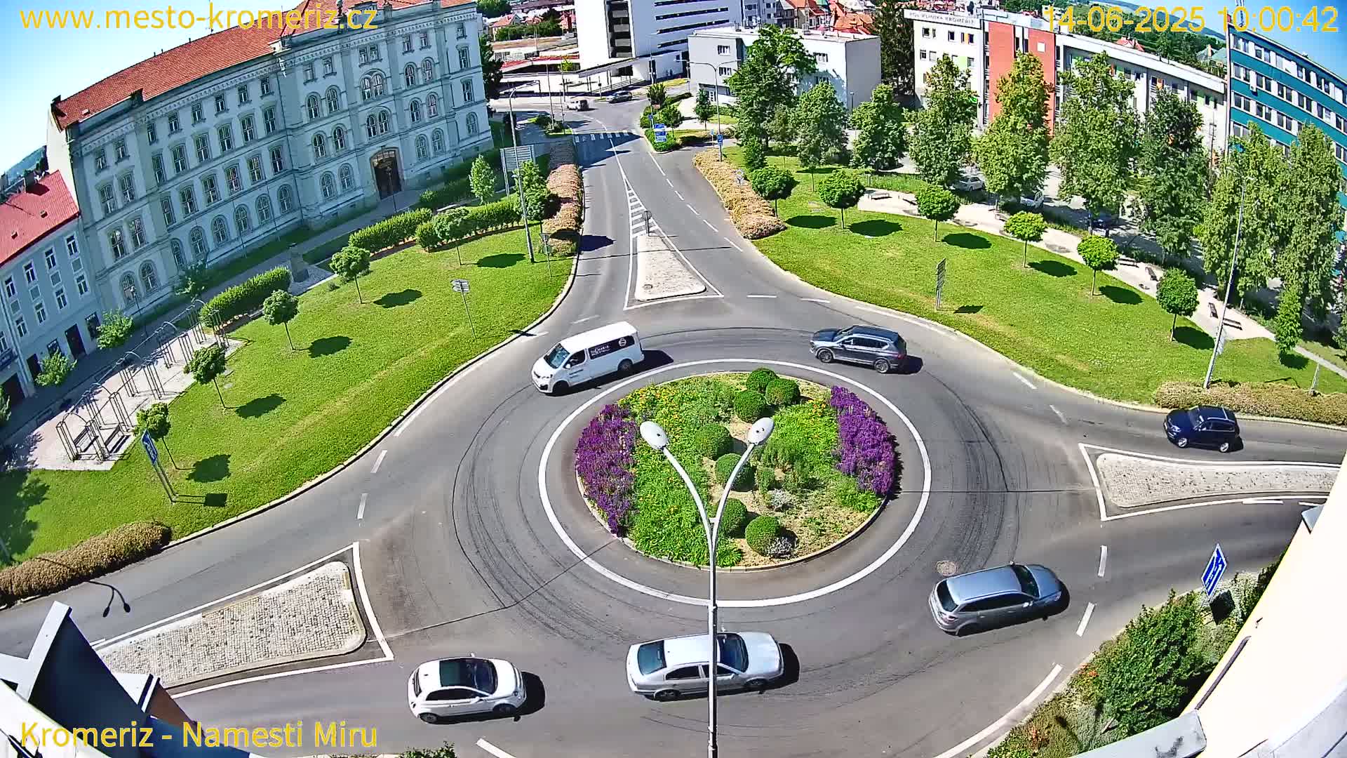An aerial view shows a sunny day overlooking a roundabout surrounded by a grassy area, buildings, and several cars navigating the circle.