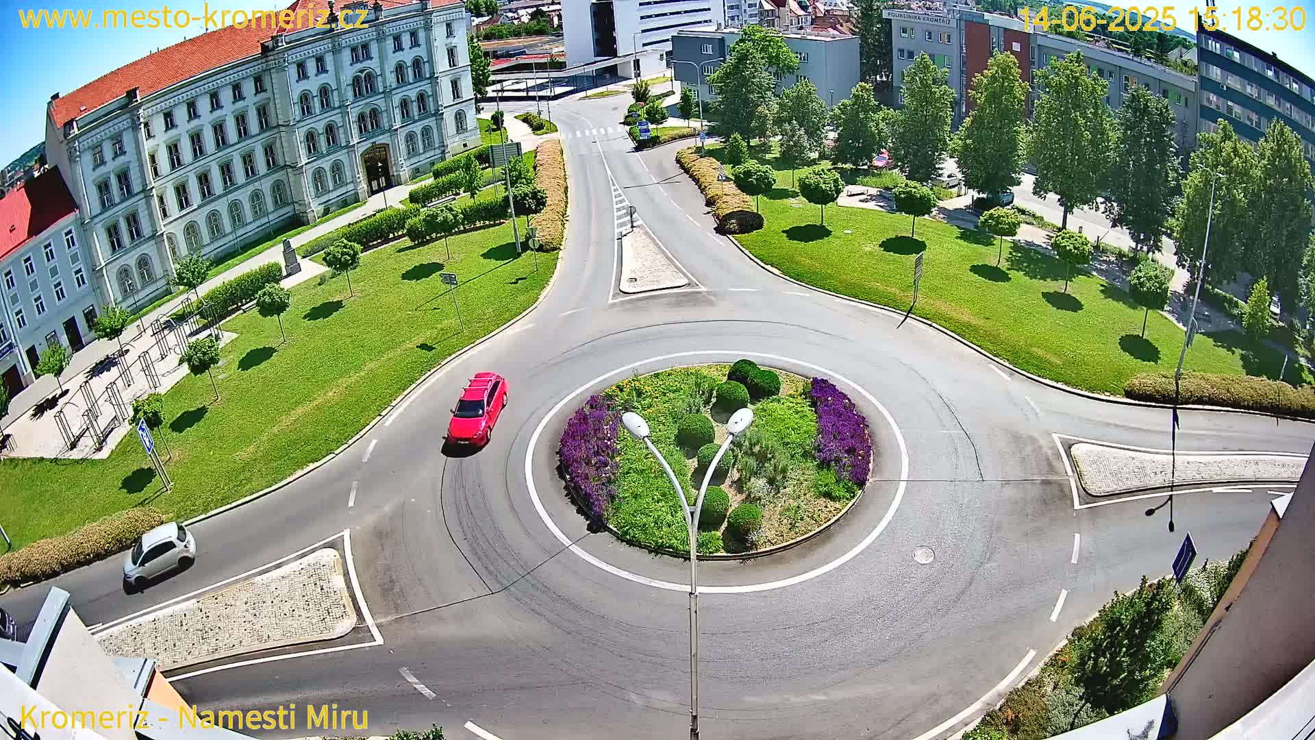 A high-angle, sunny-day view of a roundabout with two cars, surrounded by green lawns, flowering plants, and buildings.