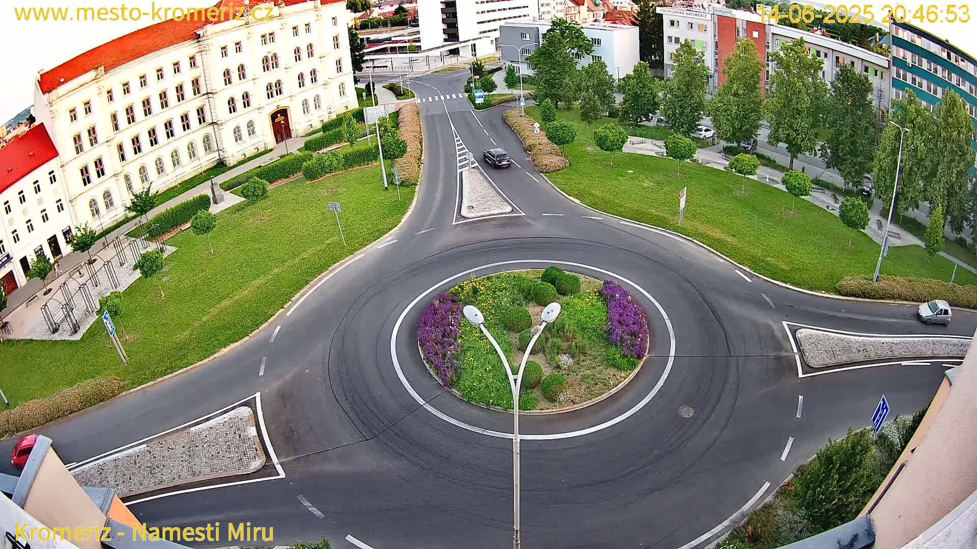 A high-angle, sunny-day view of a roundabout with two cars, surrounded by green lawns, flowering plants, and buildings.