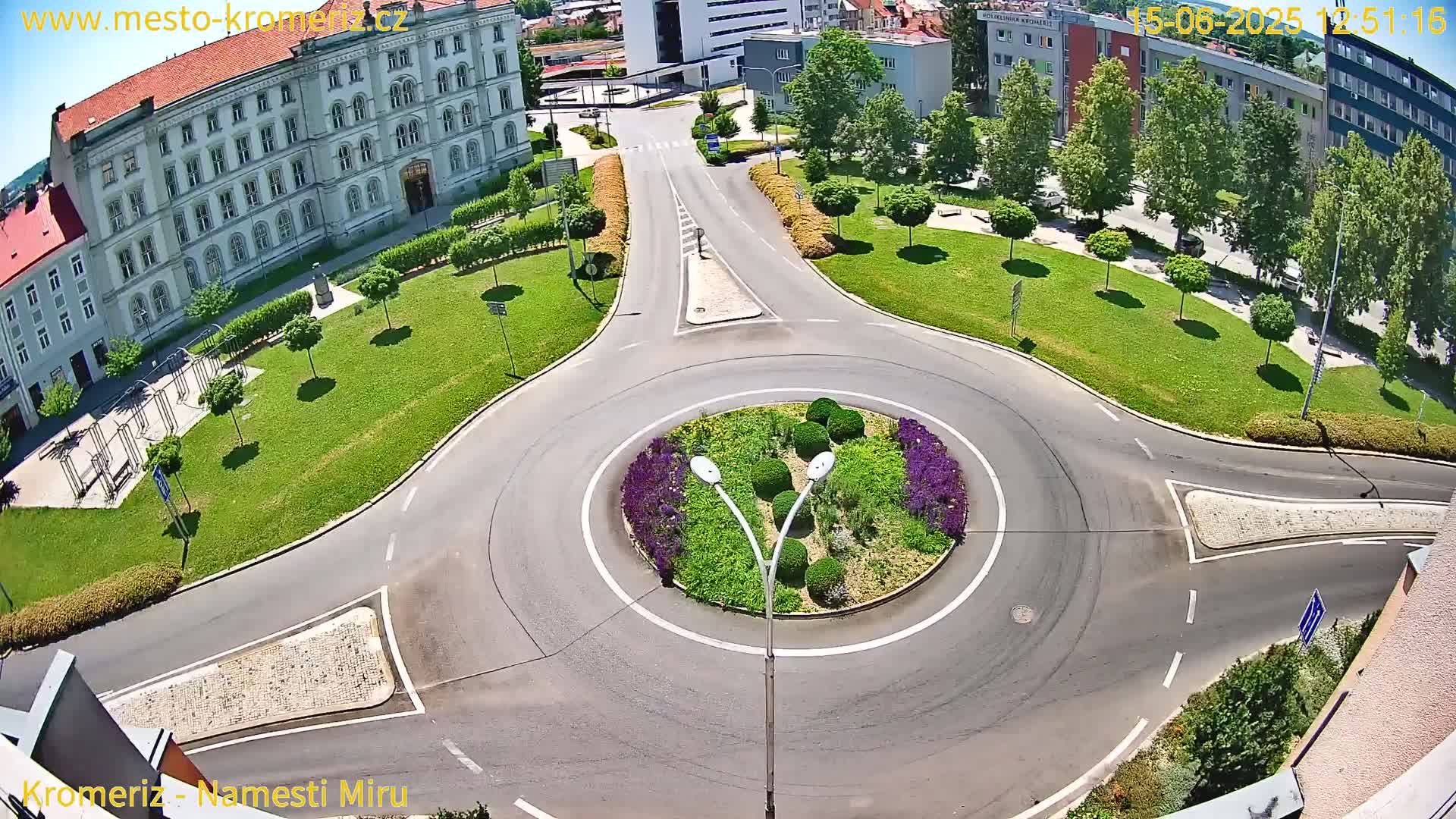 A sunny day shows a roundabout surrounded by a grassy area with trees and a large building nearby.
