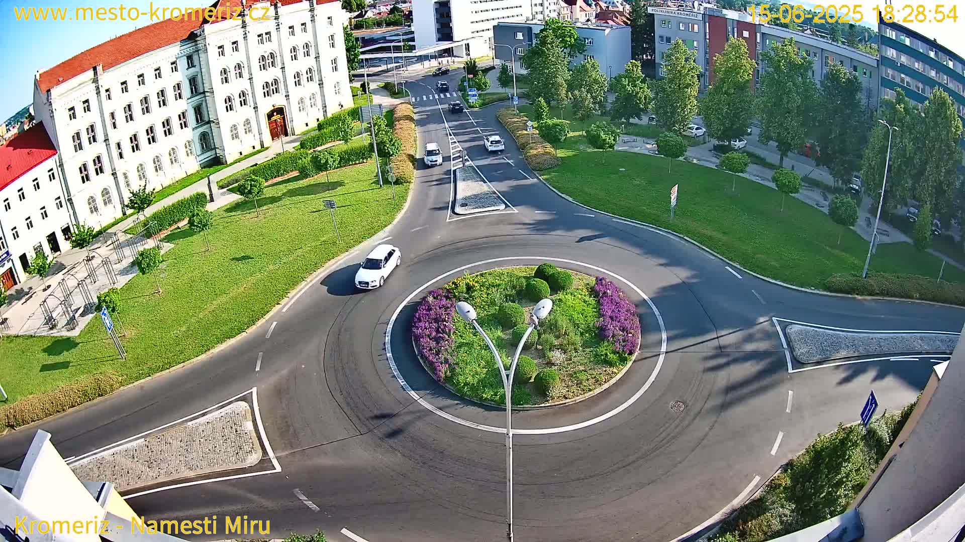 An aerial view shows a roundabout with a few cars on a sunny day, surrounded by a large building, green lawns, and trees.