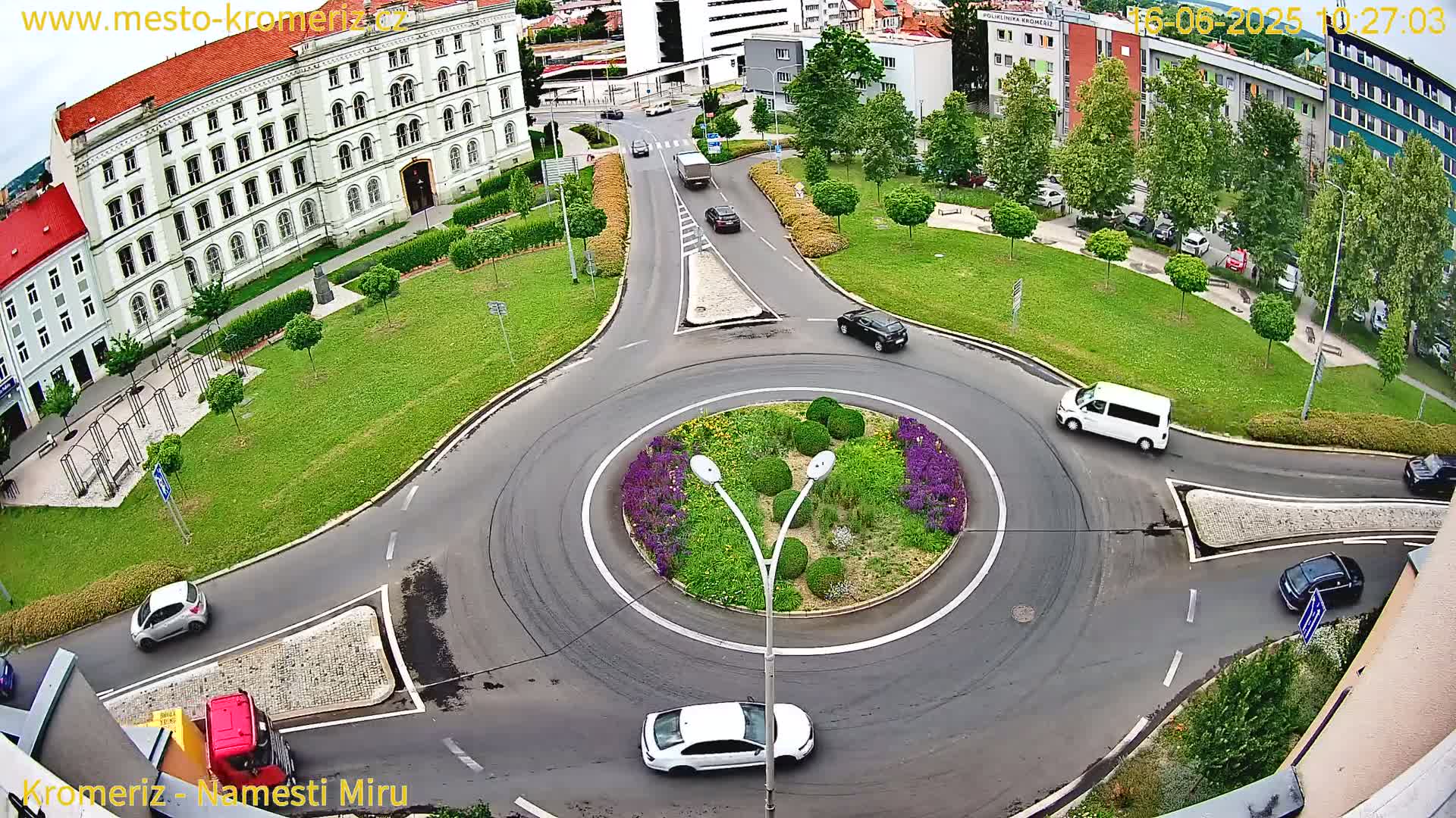 An aerial view of a roundabout with several cars and a large building in the background on a sunny day.
