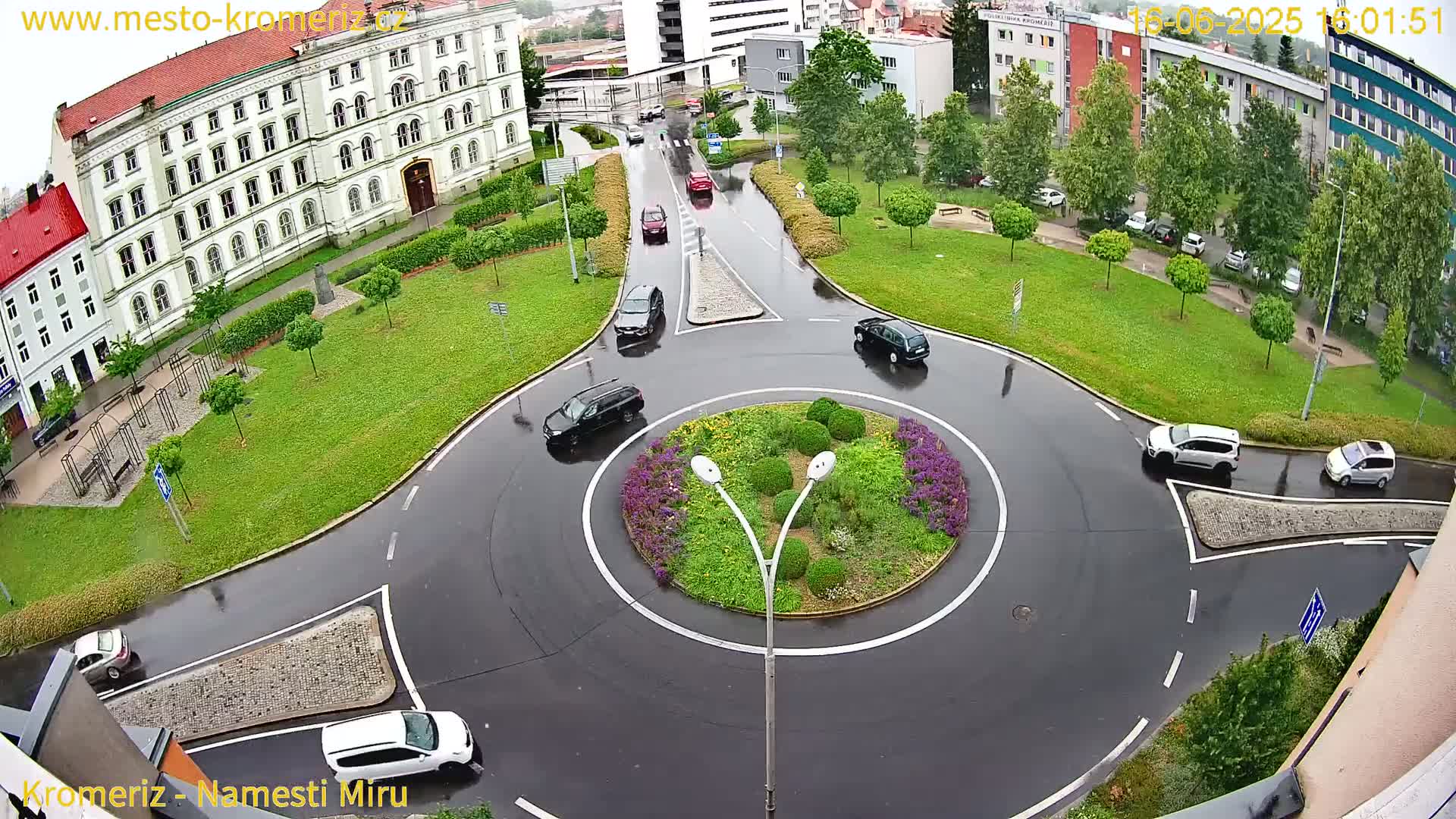 A wet, circular road intersection with several cars driving around a landscaped center island, surrounded by buildings and green space.