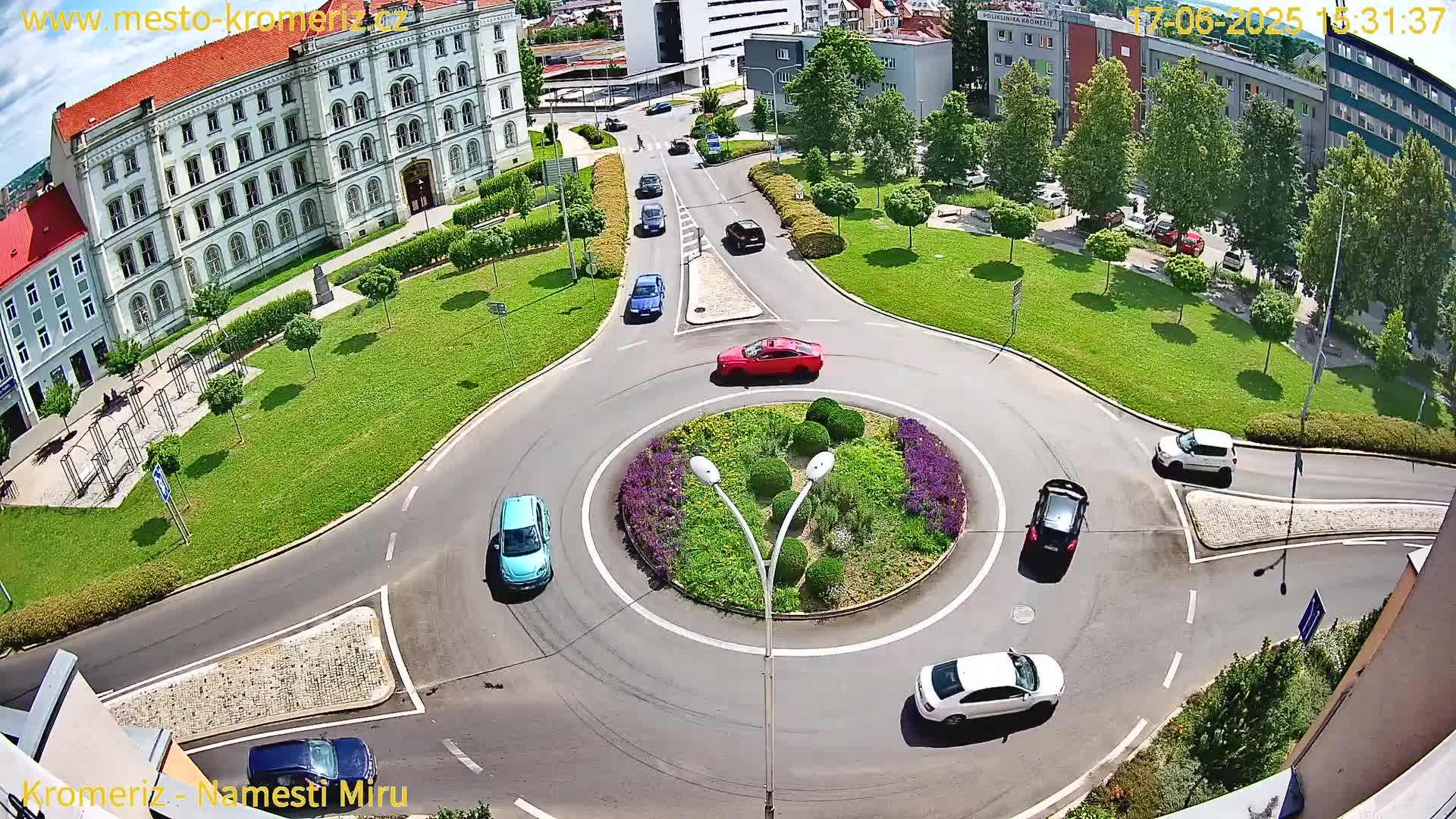 An aerial view of a roundabout surrounded by a grassy area and buildings, on a sunny day with several cars navigating the circle and nearby roads.