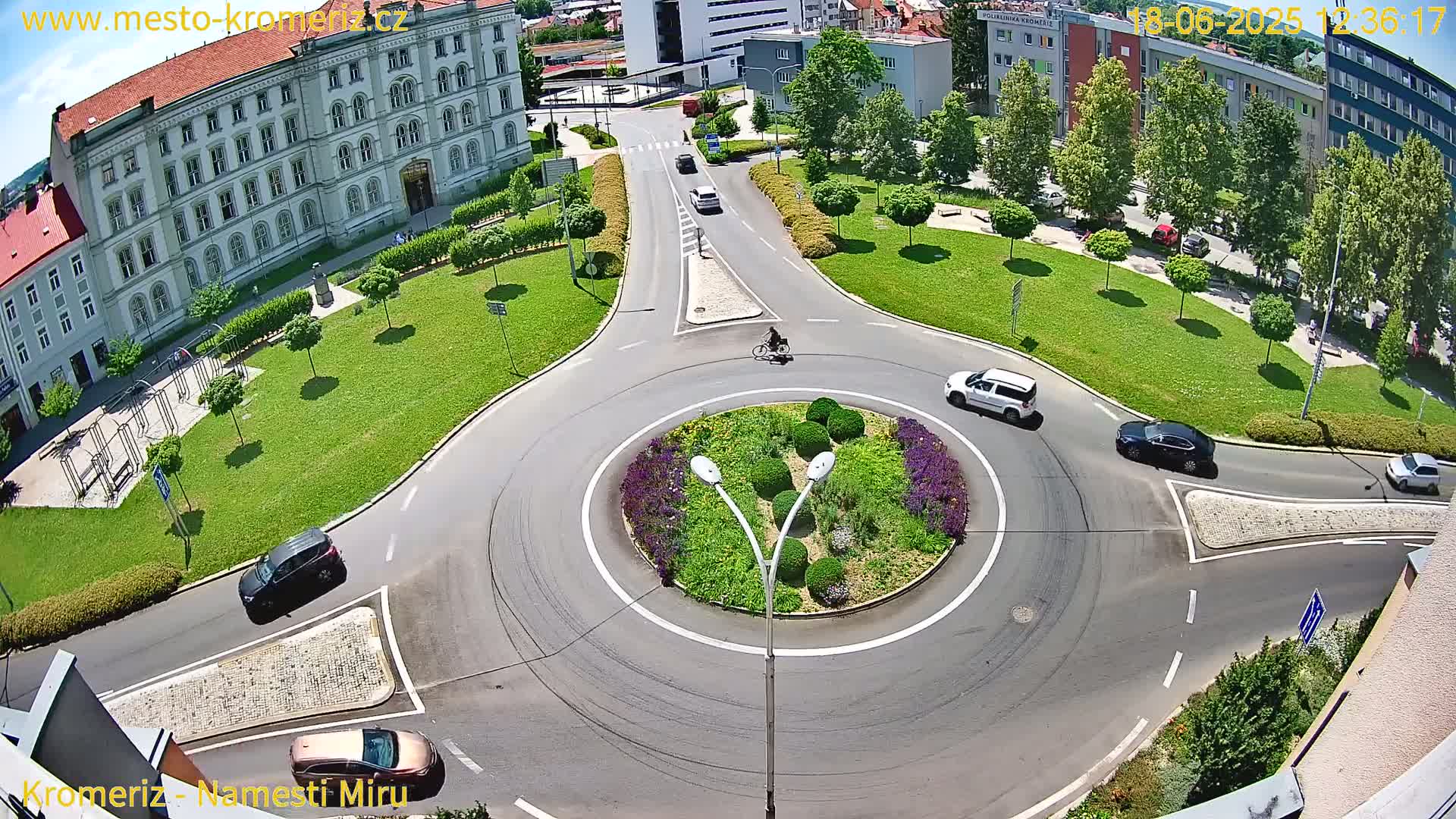 A sunny day shows a roundabout surrounded by a grassy area, buildings, and several cars and a bicycle moving through the intersection.