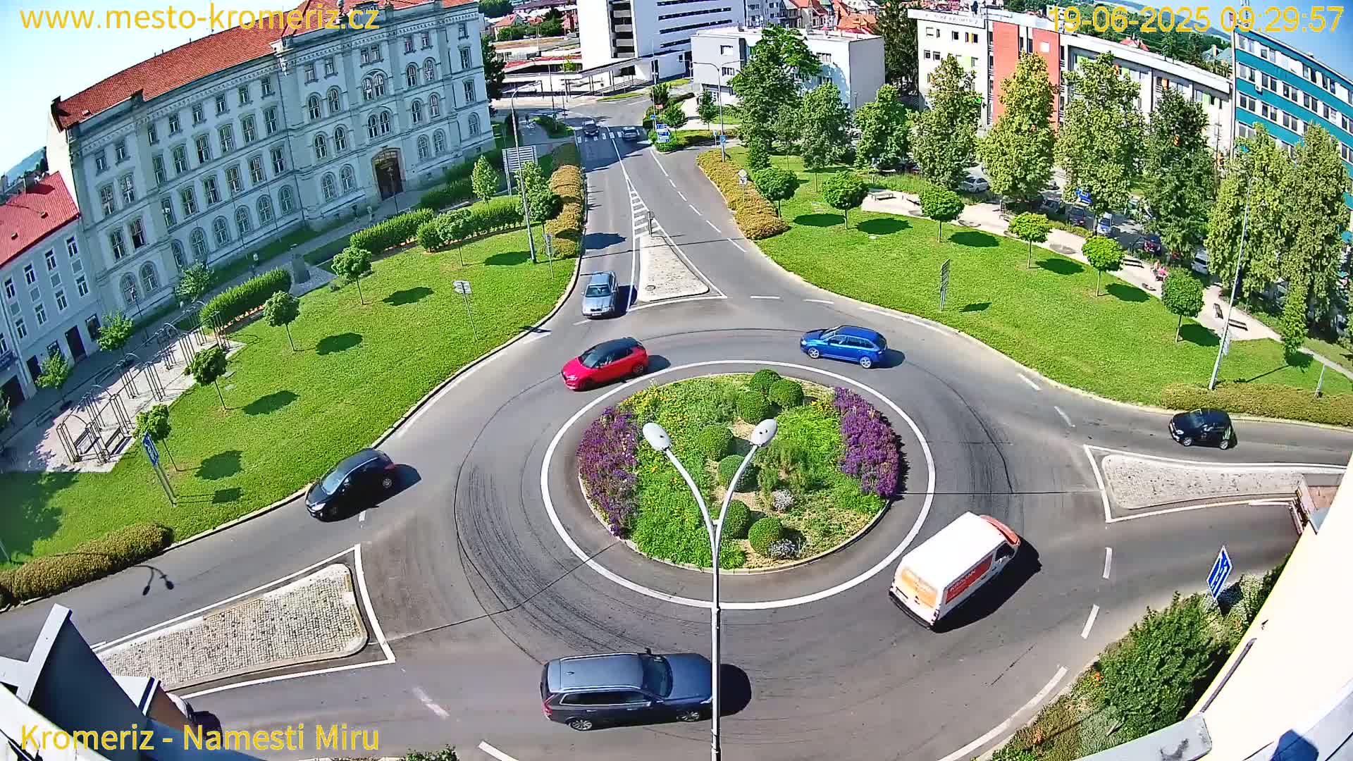 An aerial view of a roundabout surrounded by a grassy area with several cars and a van navigating it on a sunny day.