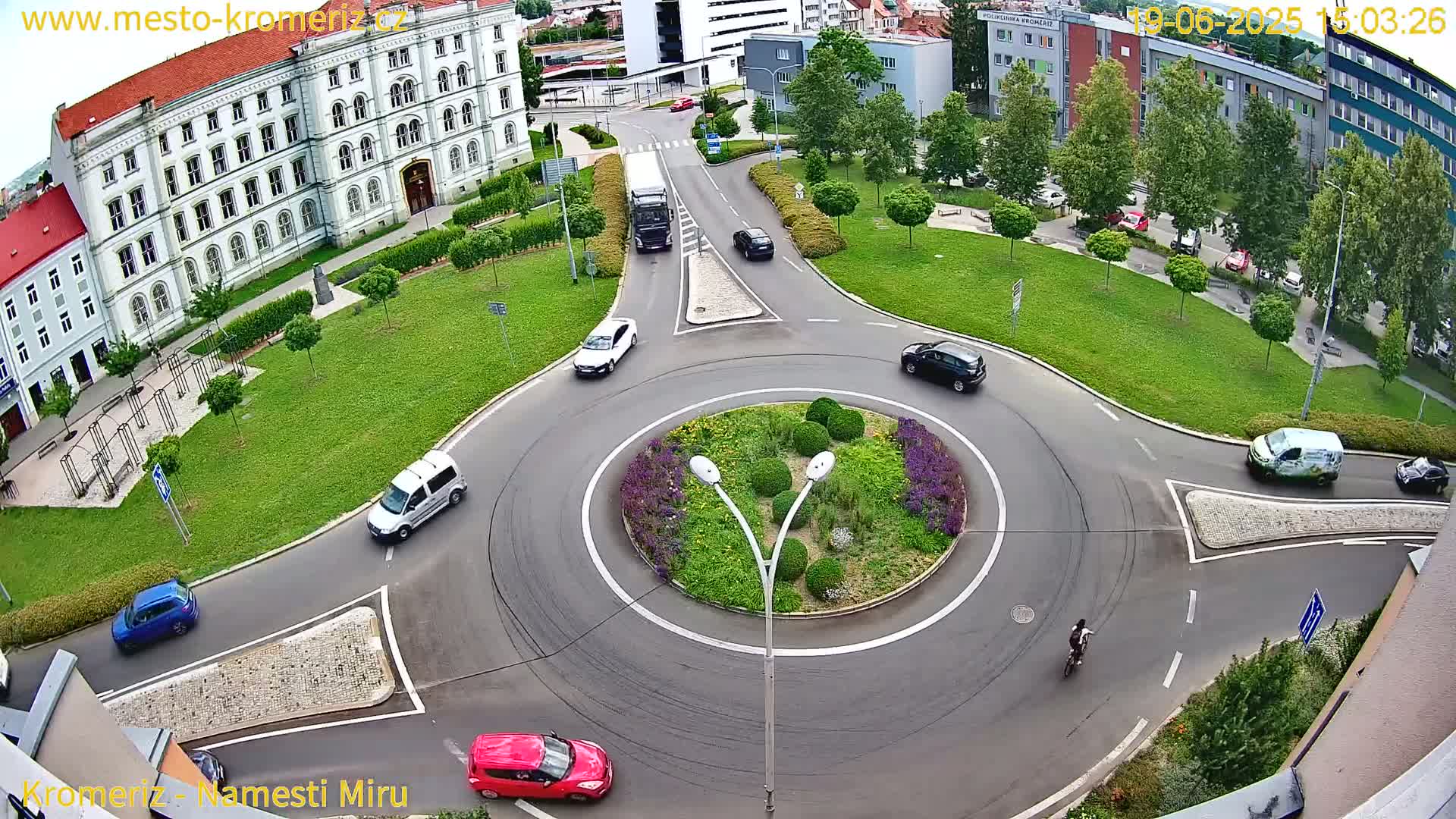 A high-angle view of a roundabout with several cars and a cyclist, surrounded by buildings and green spaces on a sunny day.