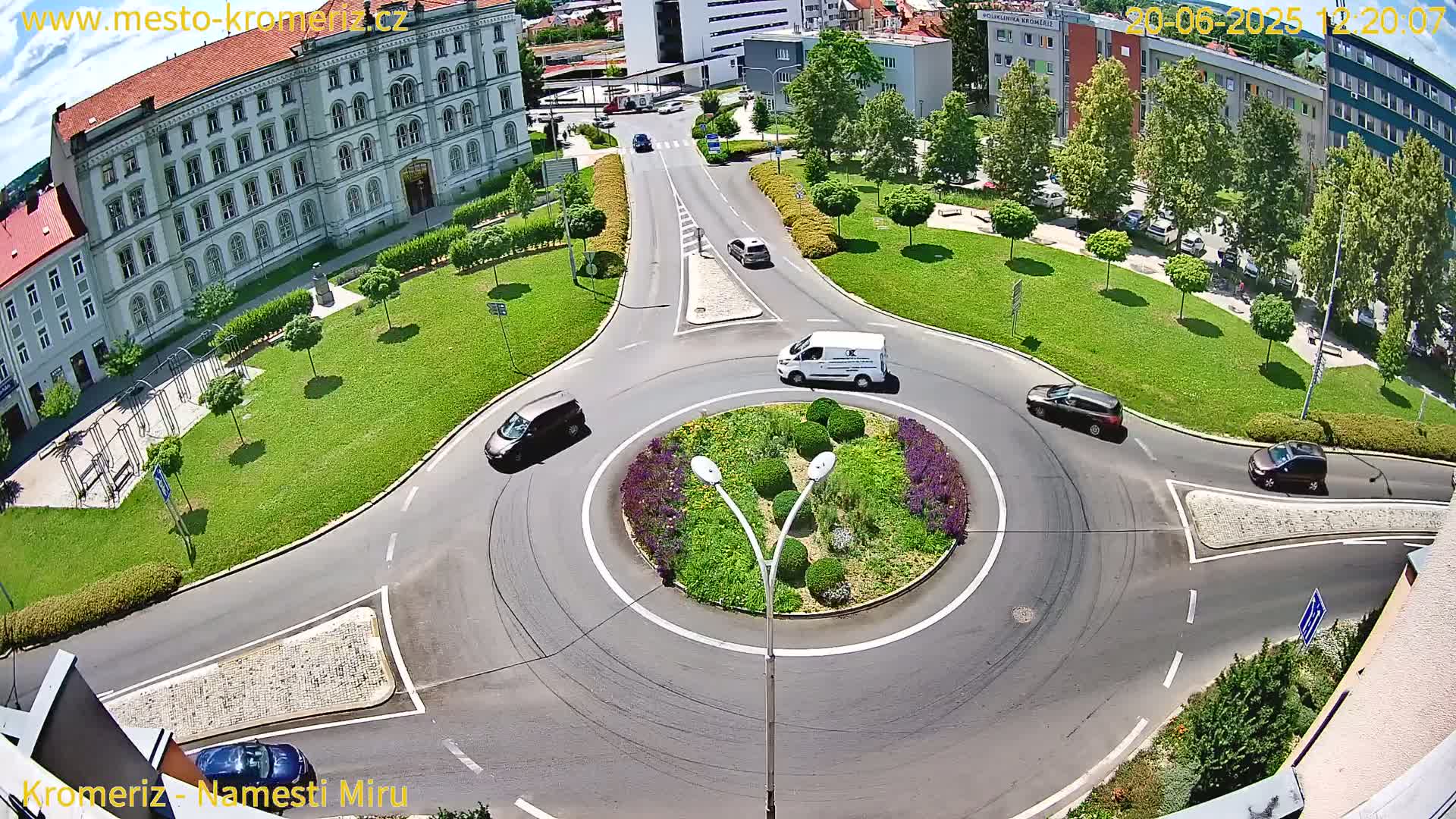 A sunny overhead view shows several cars navigating a roundabout surrounded by a landscaped median, green lawns, and buildings.