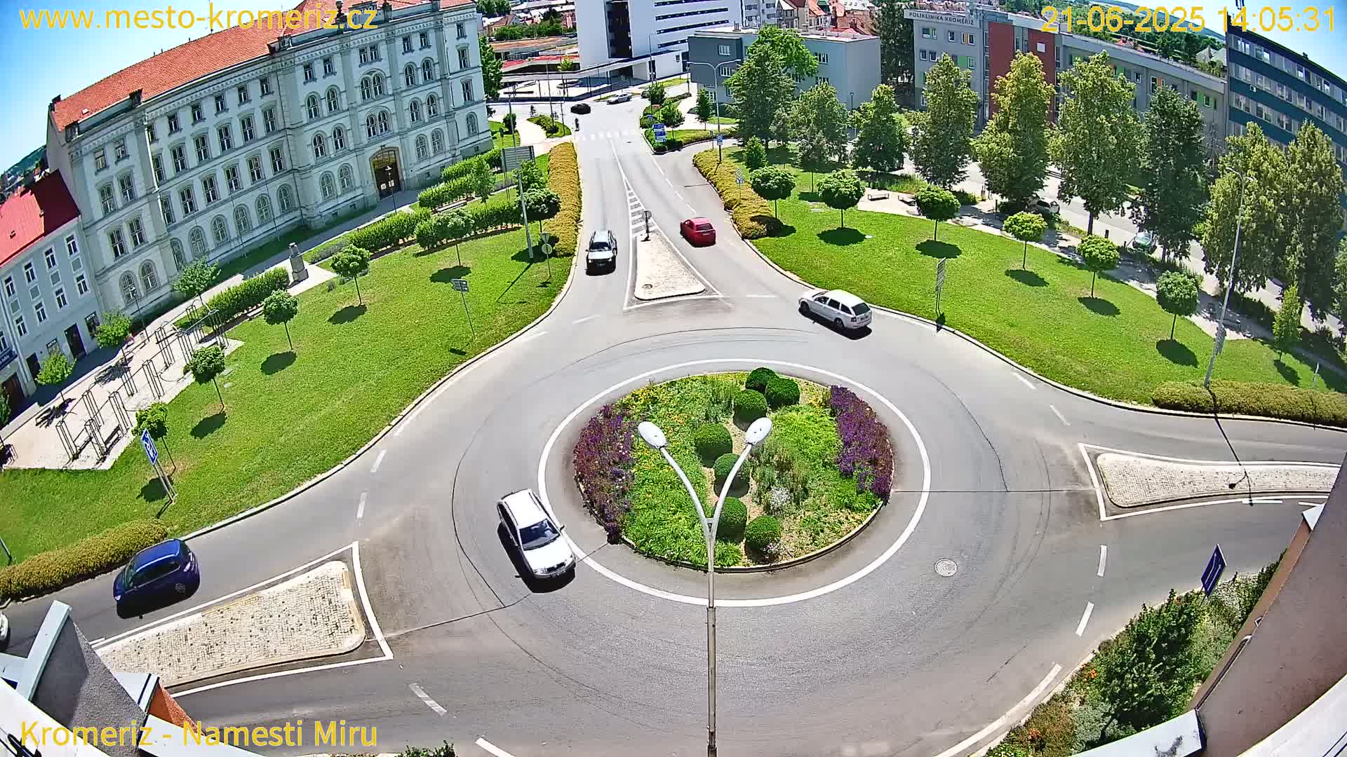 A sunny day shows a traffic circle surrounded by green lawns and buildings, with several cars navigating the roundabout.