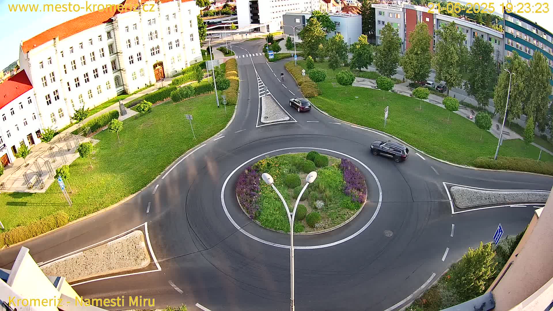 A high-angle view shows a roundabout with two cars, surrounded by buildings and green spaces, under sunny conditions.