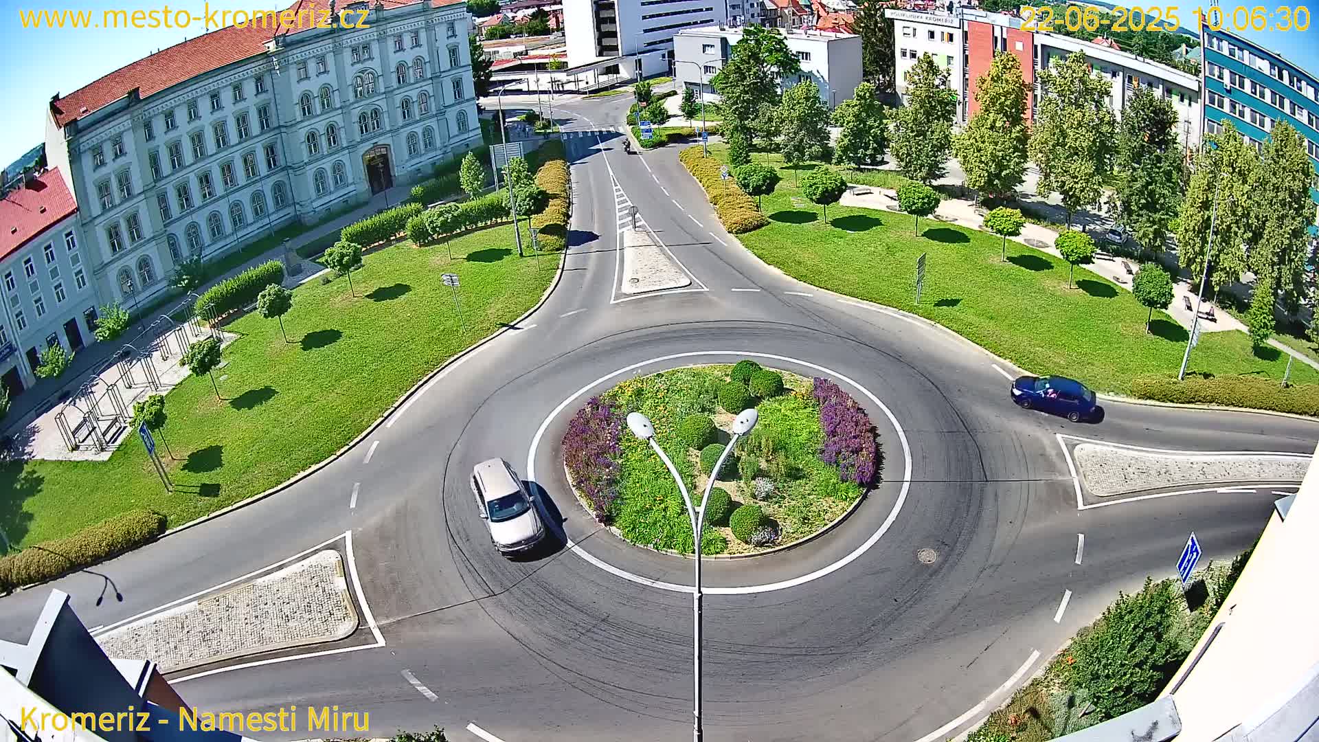 A sunny aerial view shows a roundabout with two cars, surrounded by green lawns, trees, and a large building.