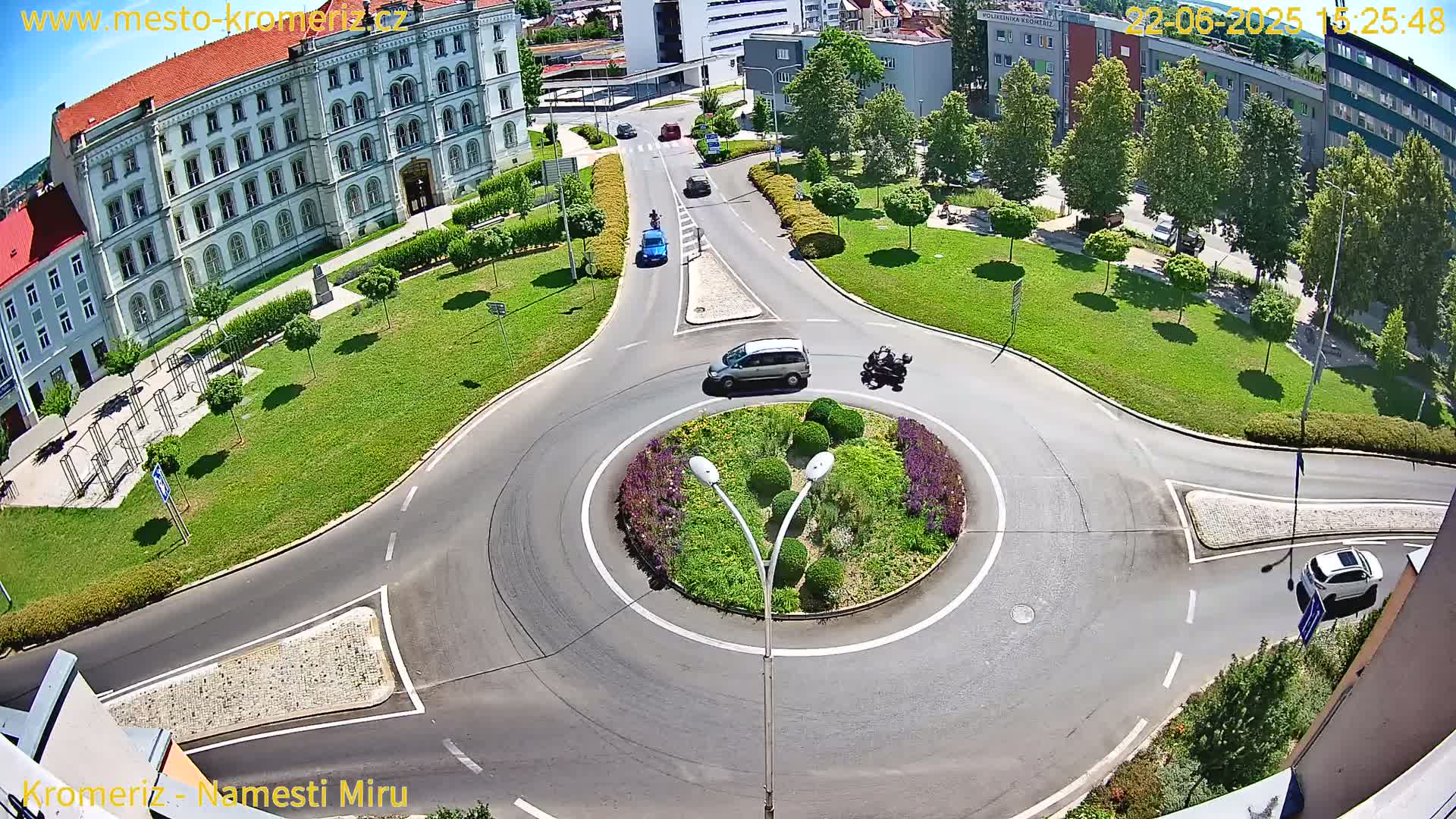 A sunny day view of a roundabout surrounded by green lawns, buildings, and several vehicles.