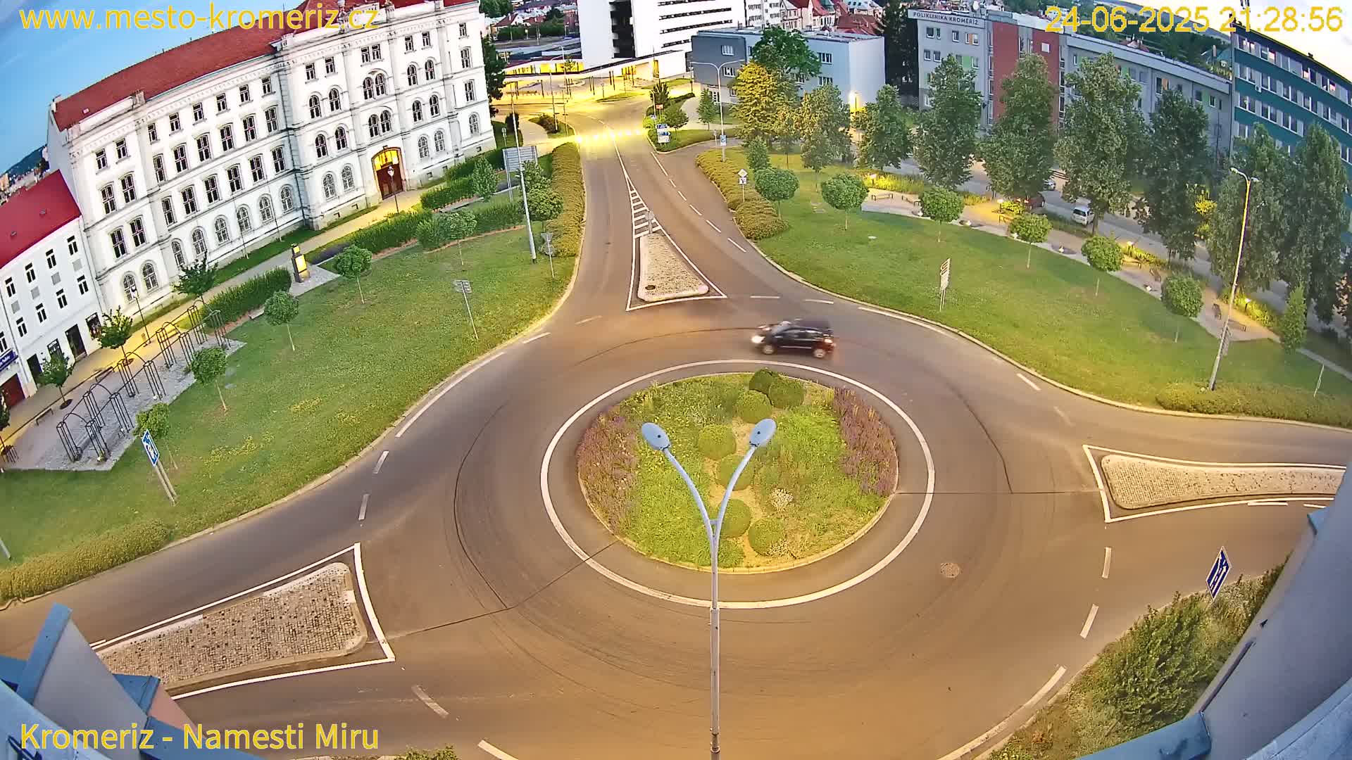 An aerial view shows a roundabout at dusk, with a single car in the circle and surrounding buildings and green spaces.