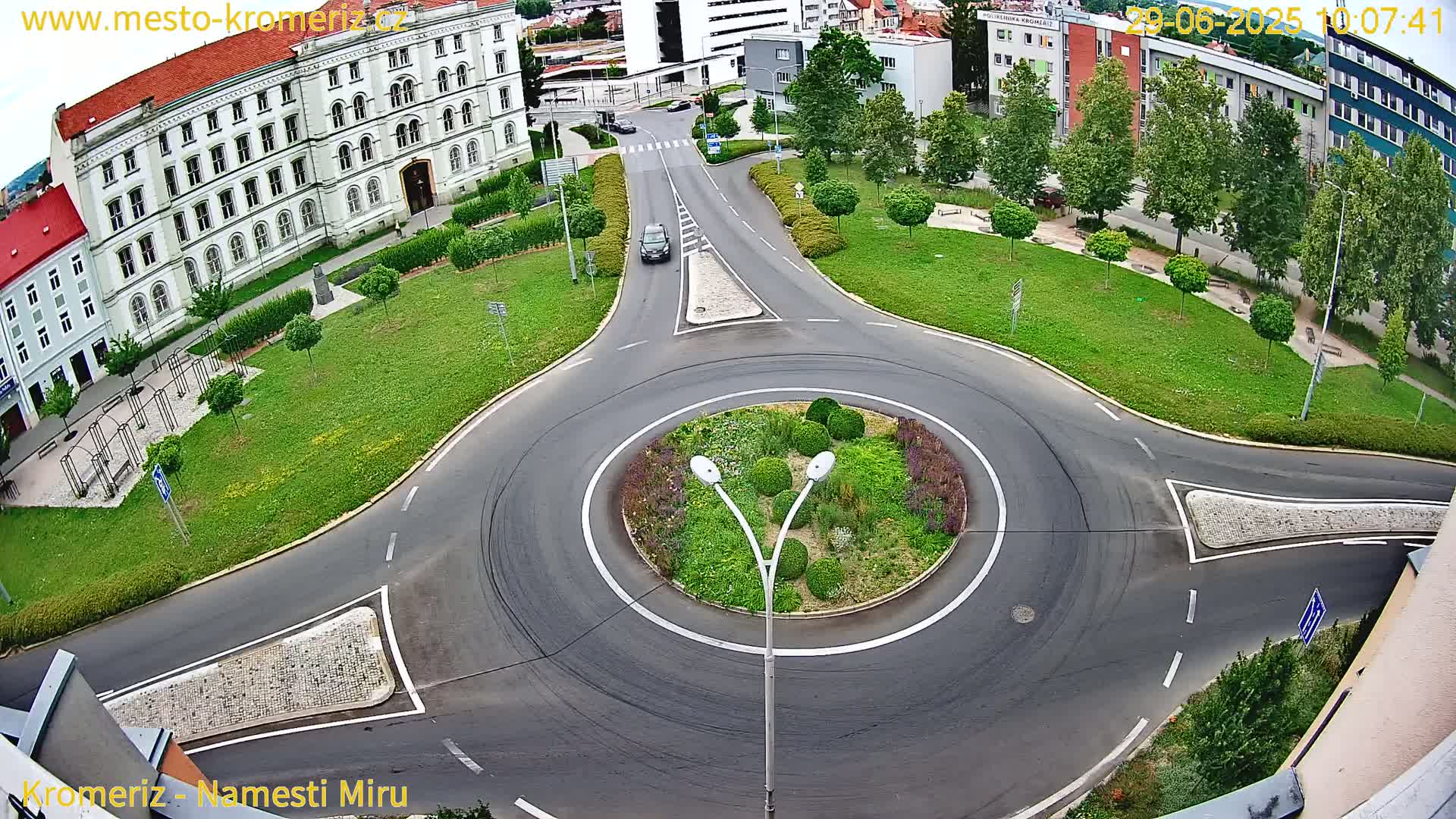 An aerial view shows a roundabout in a town square with a large building, green spaces, and a single car driving through the circle on a partly cloudy day.