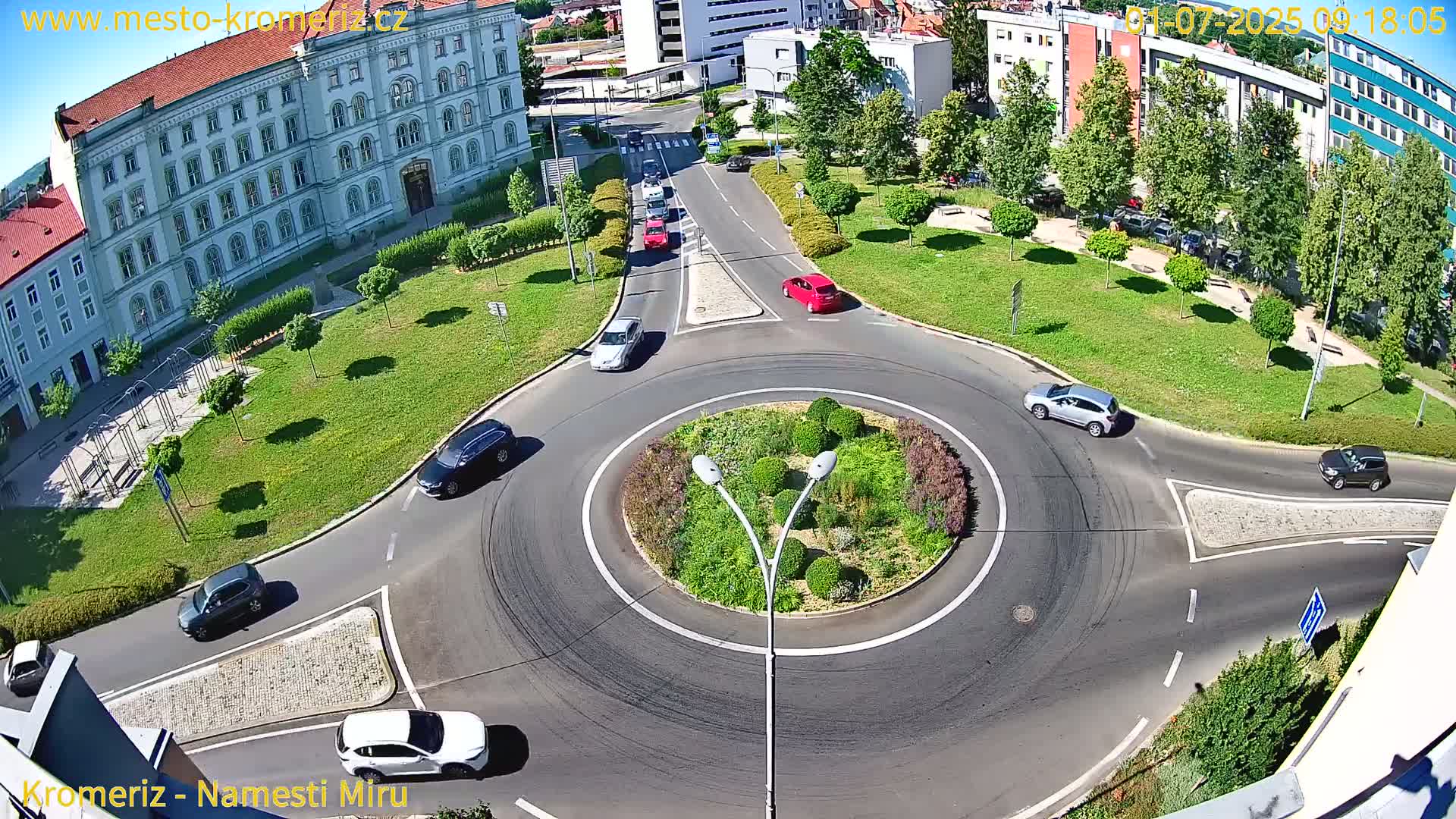 An aerial view of a roundabout with several cars circulating it, surrounded by a grassy area with trees and buildings in the background, under sunny conditions.