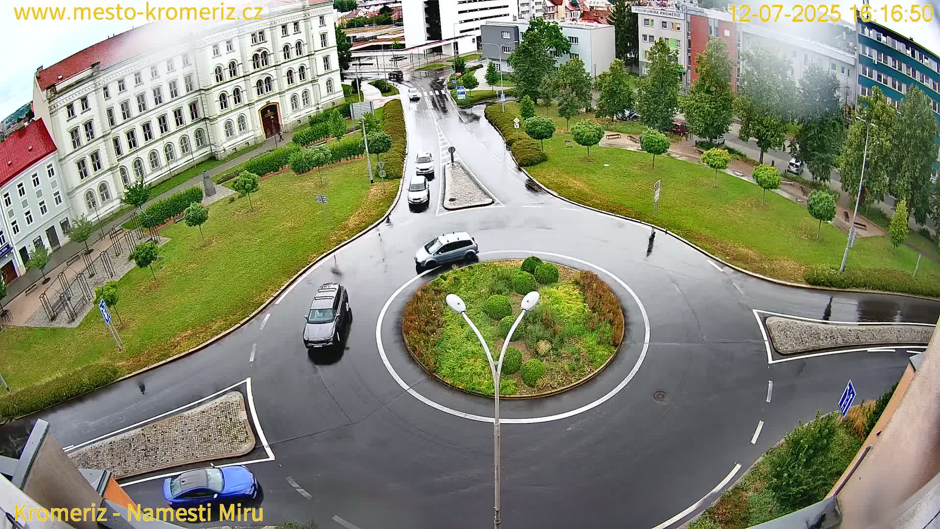 An aerial view of a roundabout with several cars circulating it, surrounded by a grassy area with trees and buildings in the background, under sunny conditions.