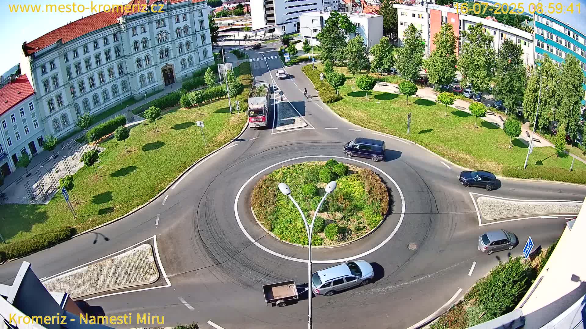 A sunny aerial view shows several cars and a truck navigating a roundabout surrounded by green lawns and buildings.