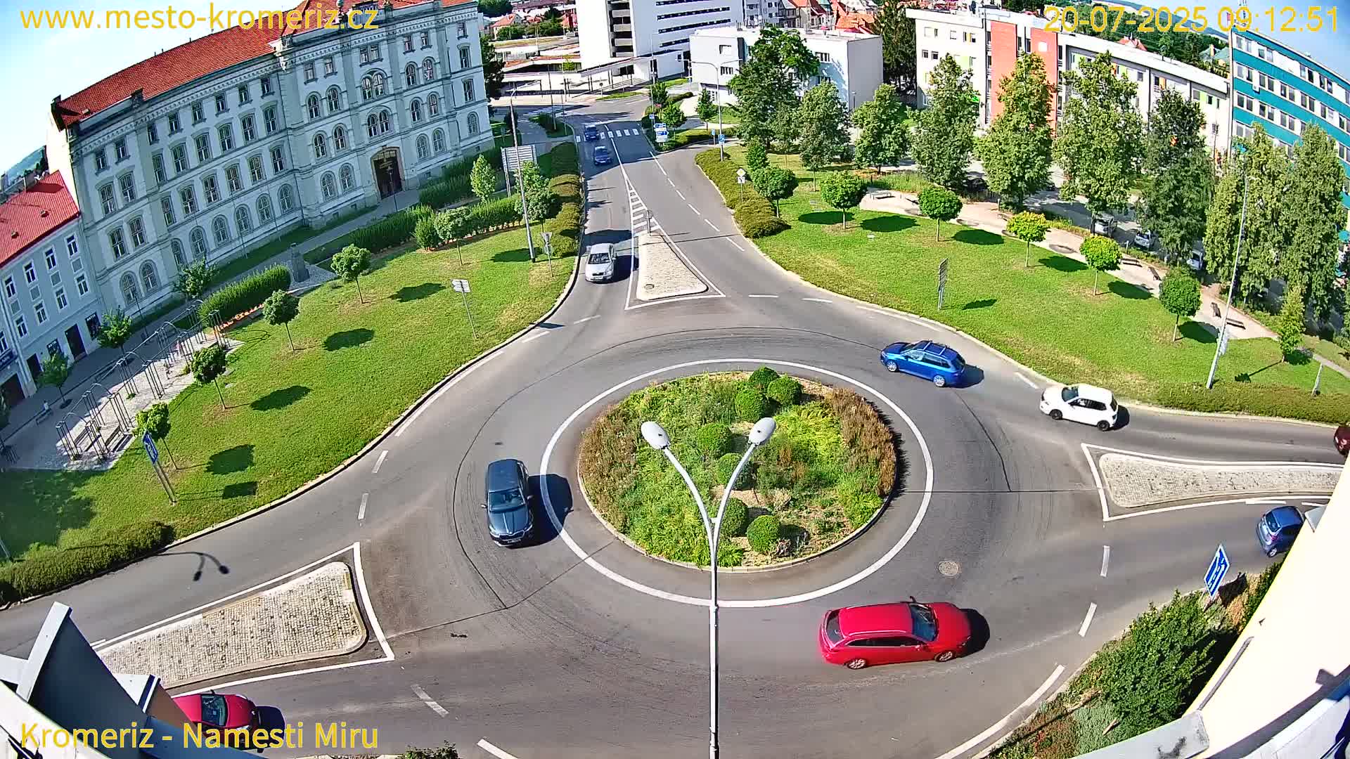A sunny day shows a roundabout with several cars driving around a landscaped center, surrounded by buildings and green spaces.