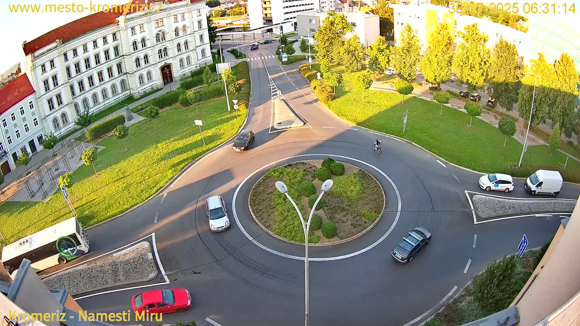 A sunny day shows a roundabout with several cars driving around a landscaped center, surrounded by buildings and green spaces.
