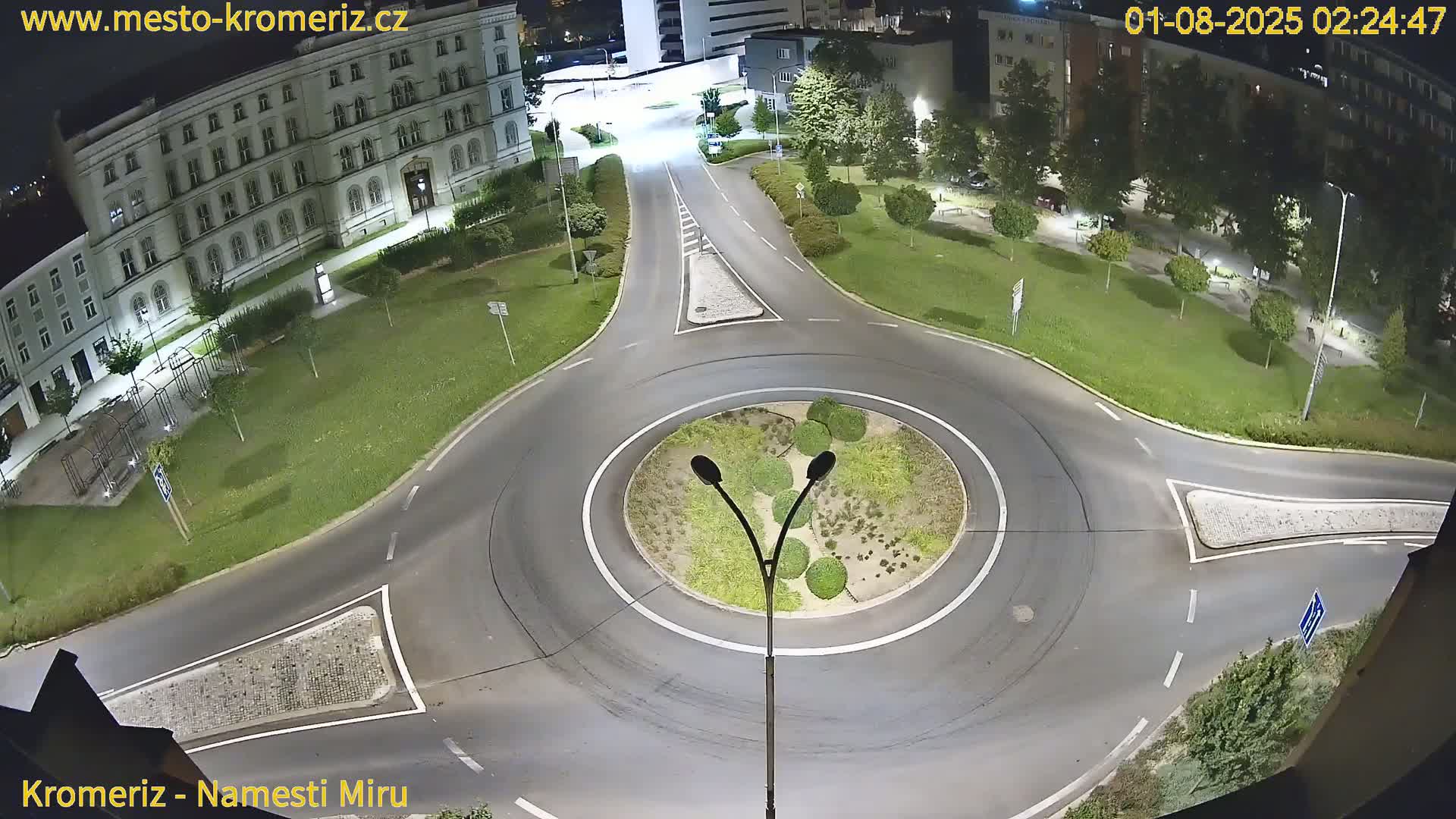 A nighttime, overhead view of a roundabout surrounded by a grassy area and buildings, illuminated by streetlights.