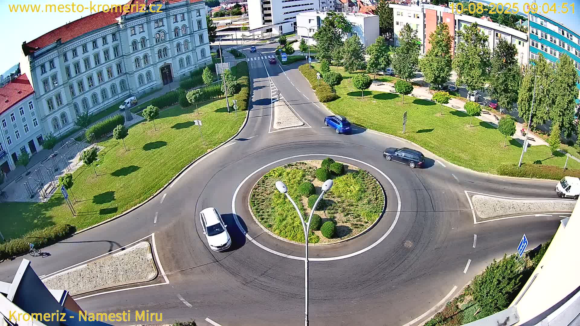 A high-angle view shows several cars navigating a roundabout on a sunny day, with a large building visible in the background.