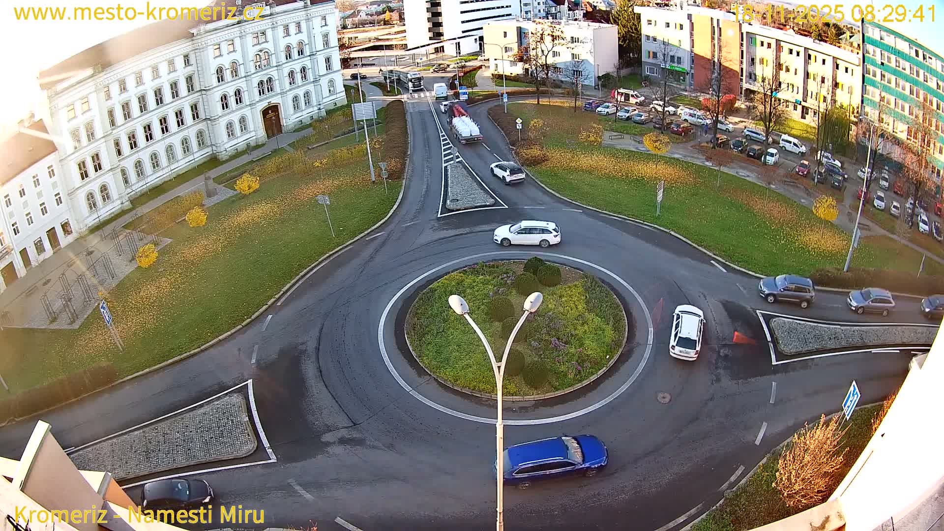 A high-angle view shows several cars navigating a roundabout on a sunny day, with a large building visible in the background.