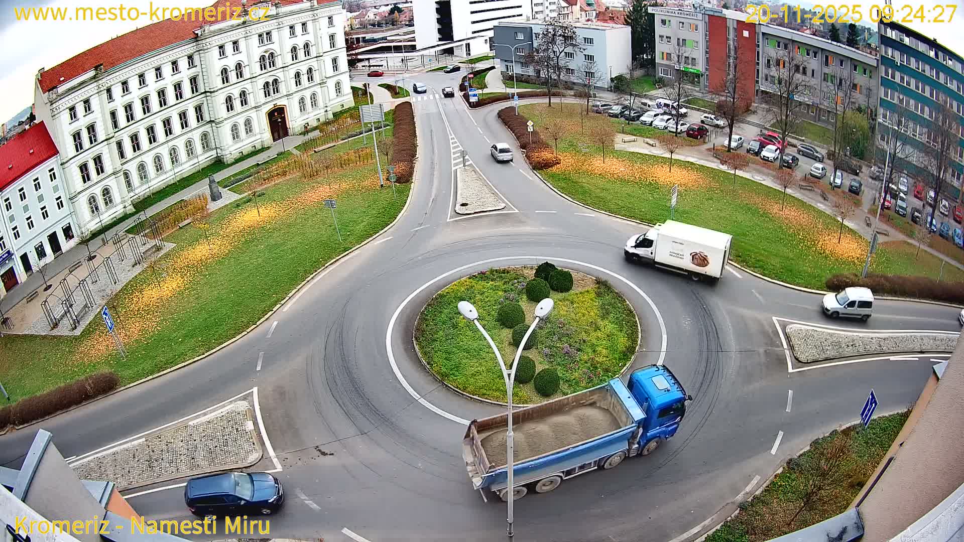 An elevated view captures a multi-lane city roundabout busy with a dump truck and a delivery van moving through it, surrounded by a mix of historical and modern buildings and green spaces with autumn foliage under an overcast sky.