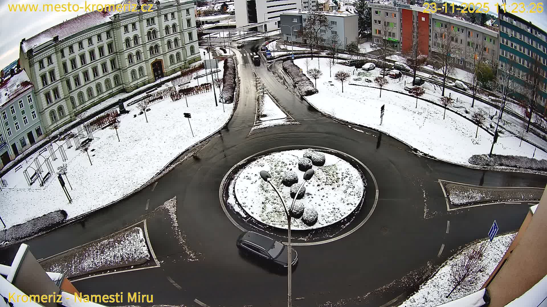 A high-angle view captures a snowy urban landscape on an overcast winter day, featuring a roundabout with a snow-covered center, wet roads, and surrounding buildings and parks dusted with snow.