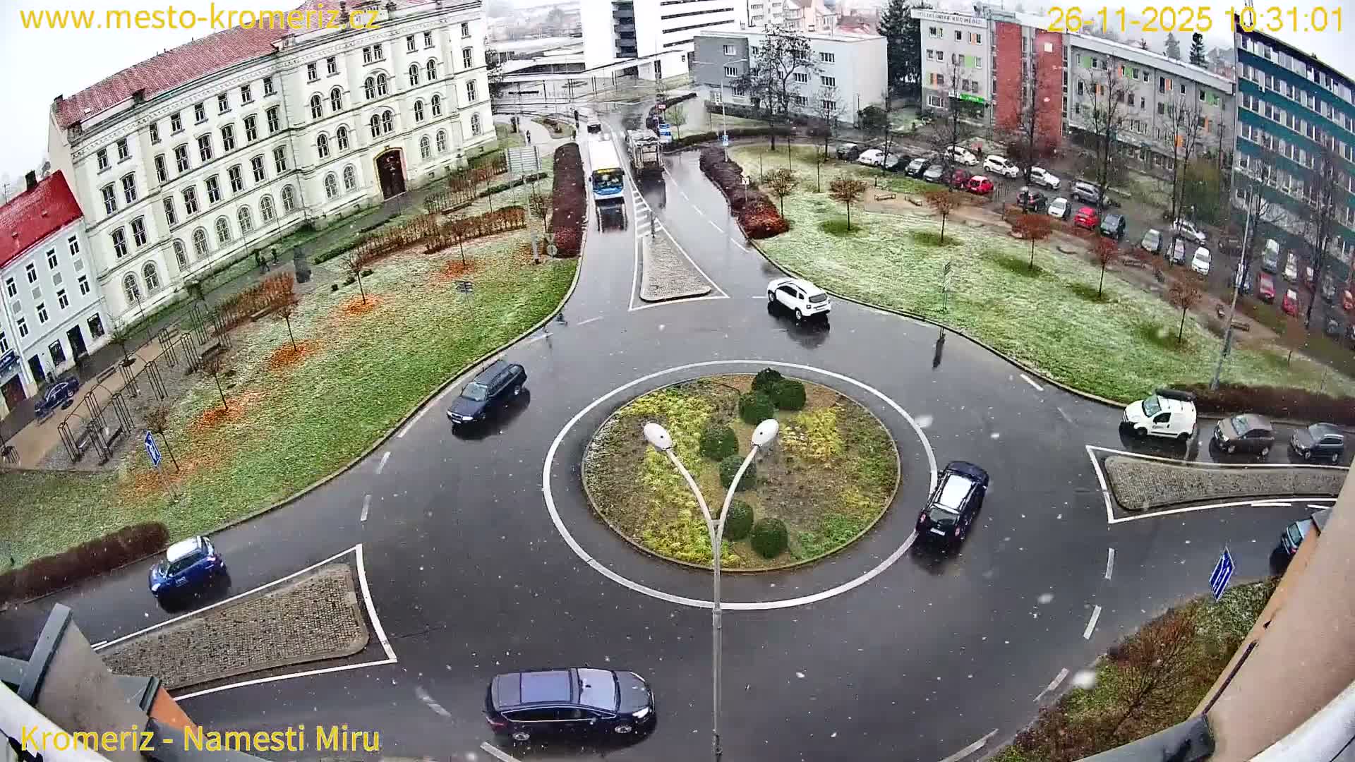 An elevated view captures a bustling urban roundabout with wet roads and several vehicles, surrounded by multi-story buildings and snow-dusted green spaces, as light snow falls on an overcast day.