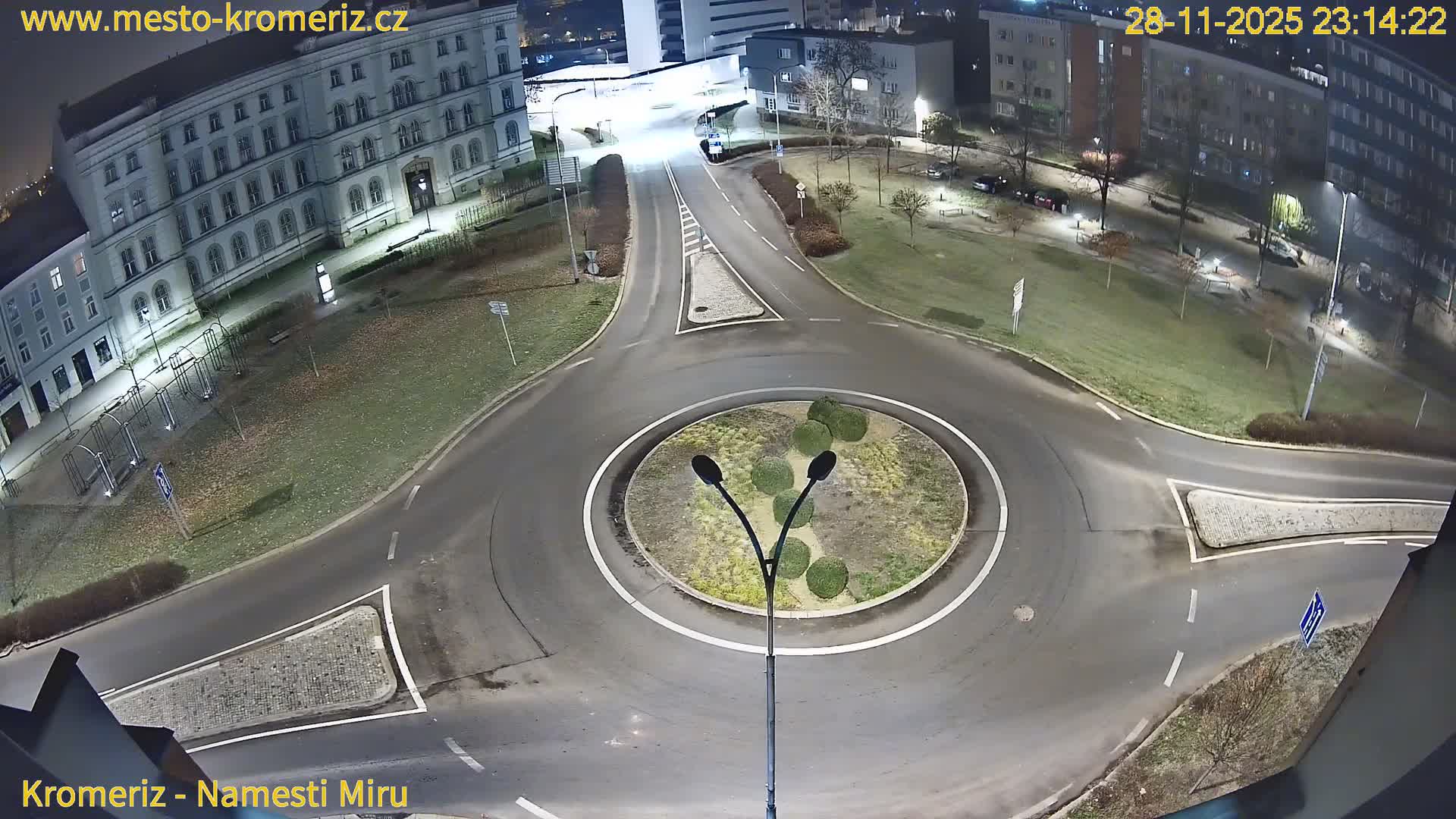 A clear night view from above shows a well-lit city roundabout surrounded by multi-story buildings, grassy areas with sparse trees, and illuminated streets with a few distant cars.