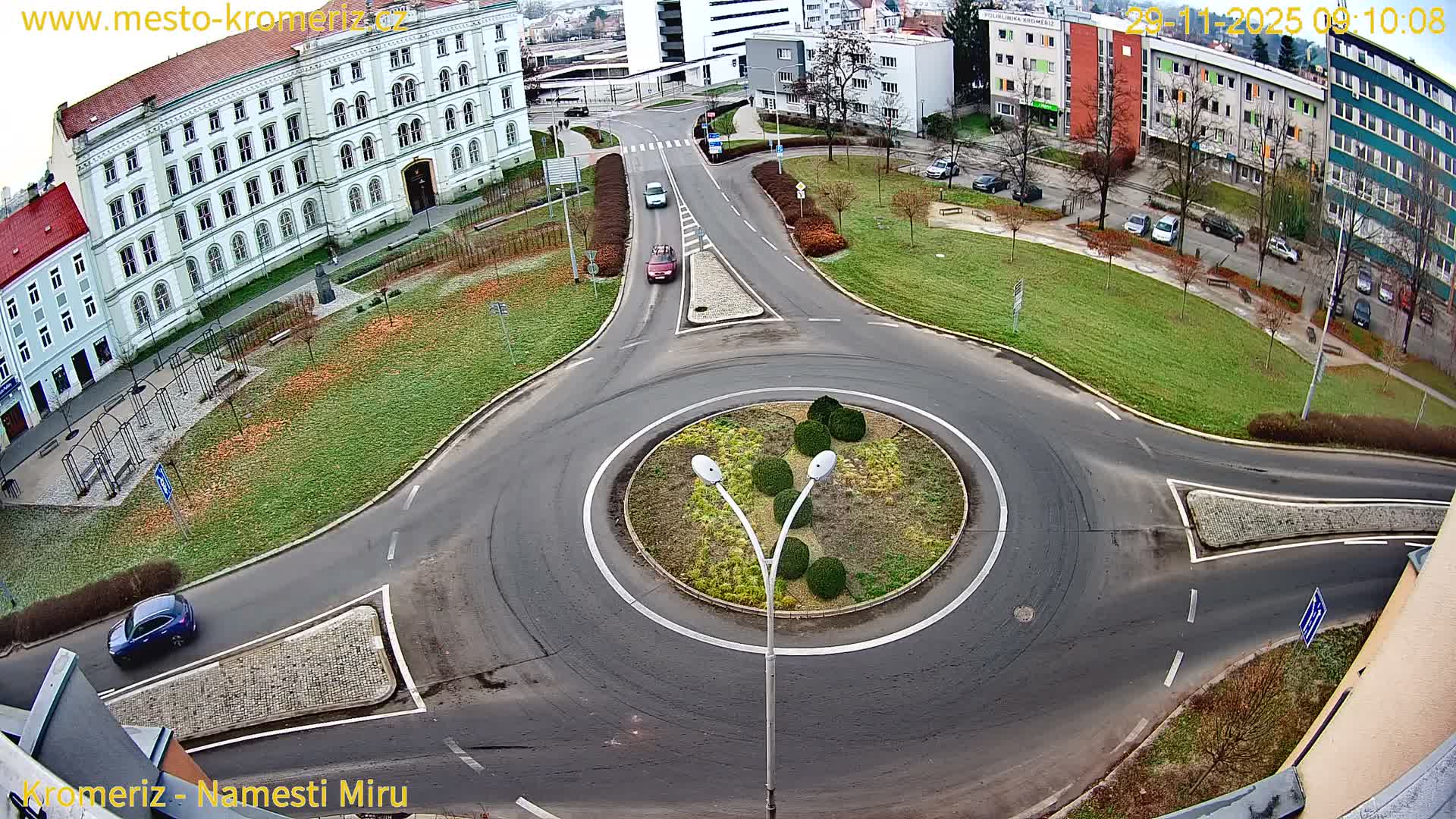 An aerial view captures a city roundabout with cars driving on a network of damp roads, flanked by grassy areas with scattered autumn leaves, bare trees, and a mix of traditional and modern buildings under an overcast sky.