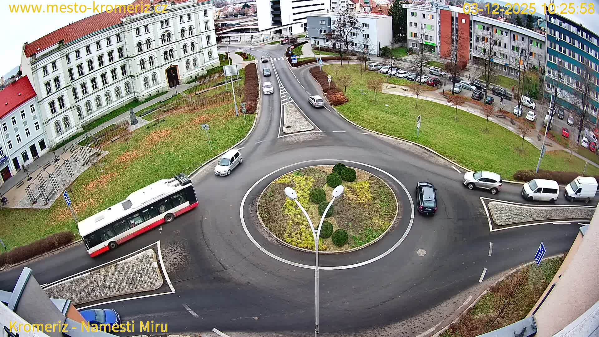 An elevated perspective captures a bustling urban roundabout with a white and red bus and several cars navigating wet asphalt roads, surrounded by buildings and sparse autumn foliage under an overcast sky.