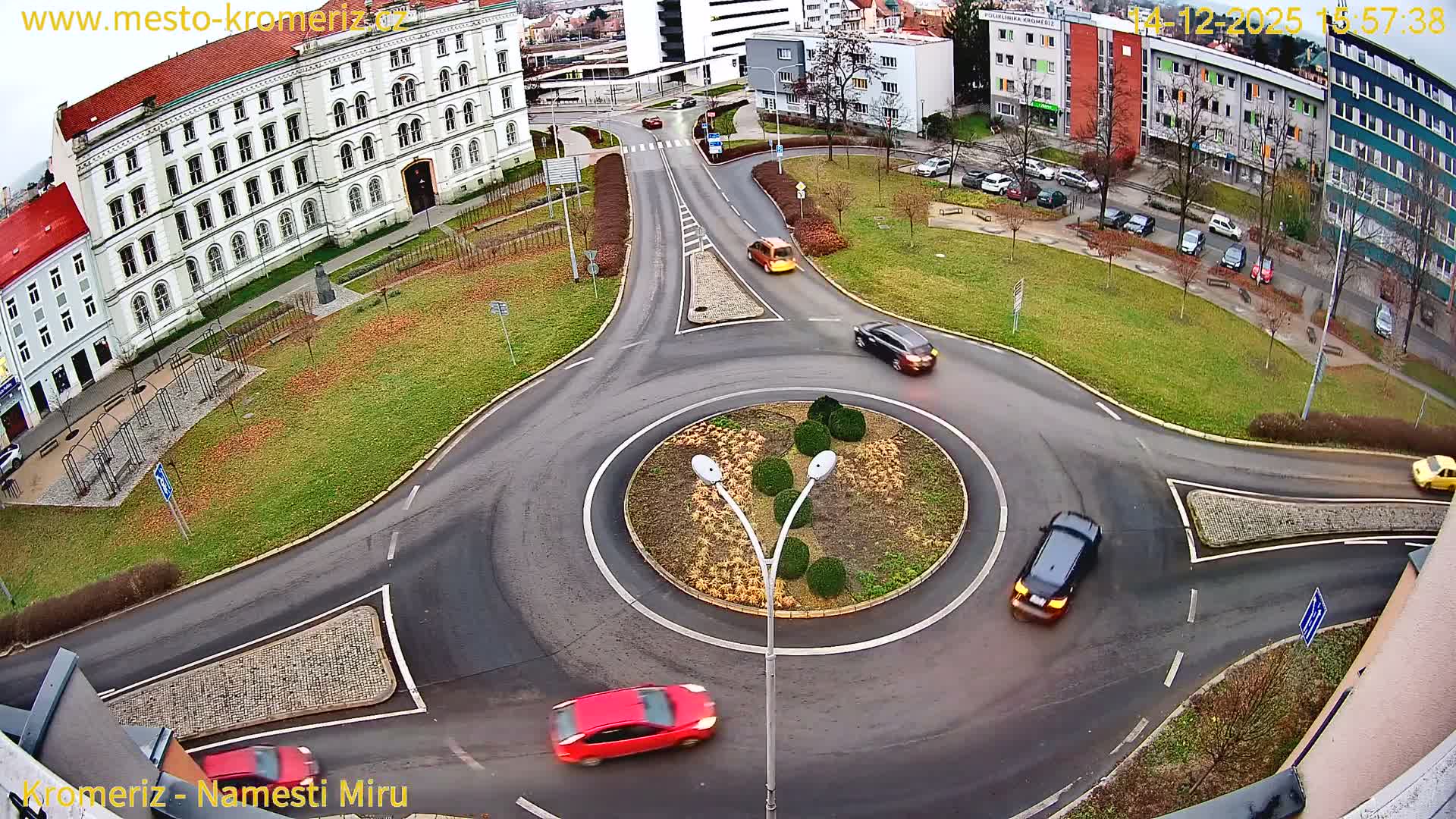An elevated perspective captures a bustling urban roundabout with a white and red bus and several cars navigating wet asphalt roads, surrounded by buildings and sparse autumn foliage under an overcast sky.