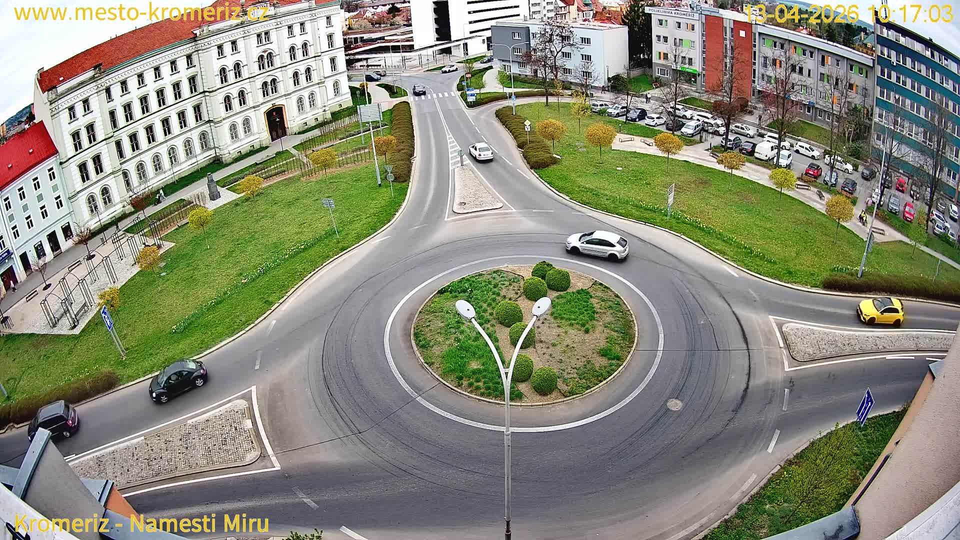 An elevated perspective captures a bustling urban roundabout with a white and red bus and several cars navigating wet asphalt roads, surrounded by buildings and sparse autumn foliage under an overcast sky.
