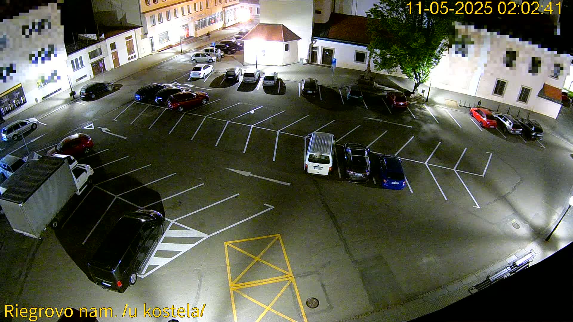 A nighttime aerial view of a mostly empty parking lot with several cars parked, surrounded by buildings under artificial lighting.