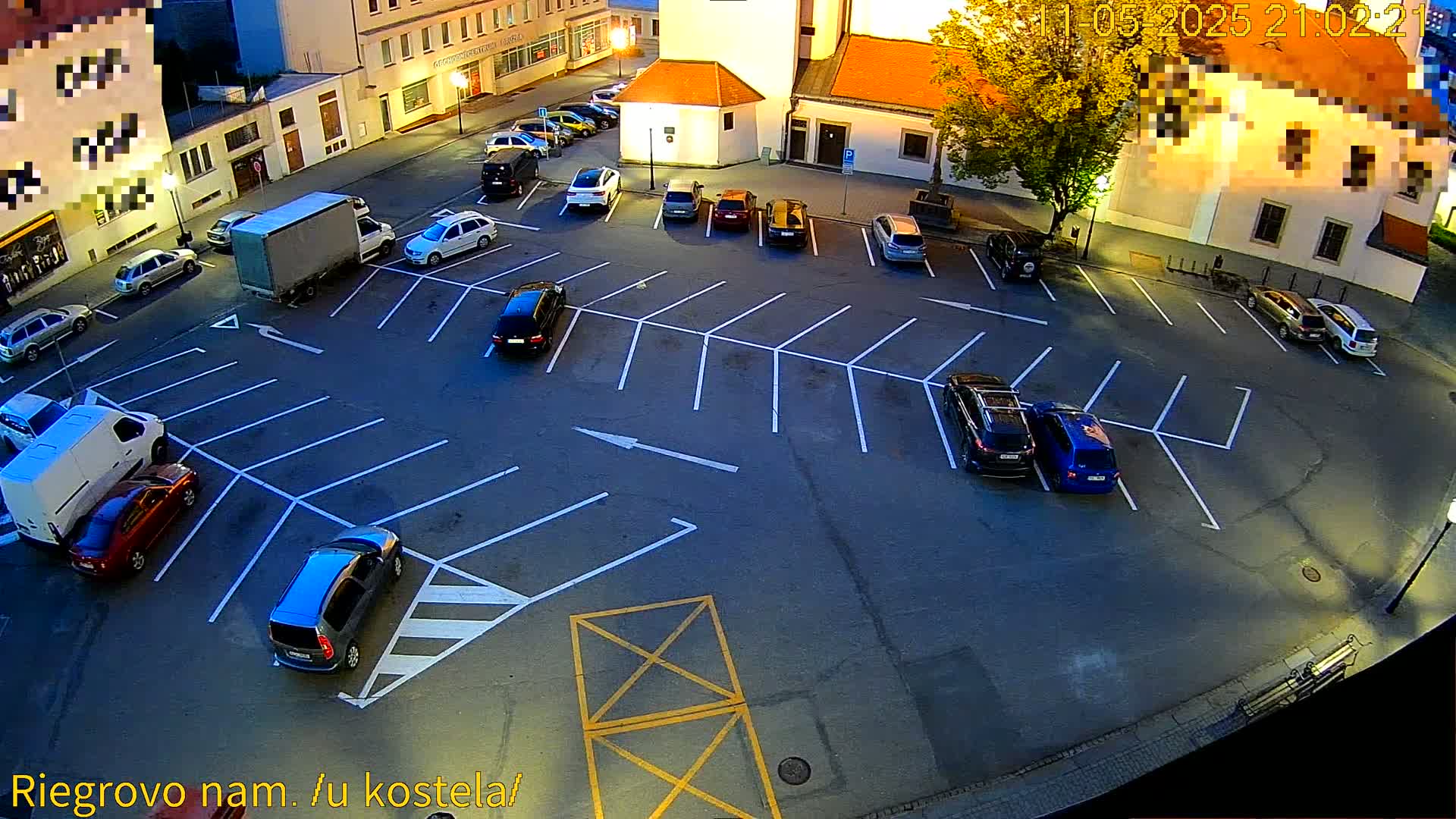 An empty asphalt parking lot at night, surrounded by buildings, with several cars parked in the spaces.