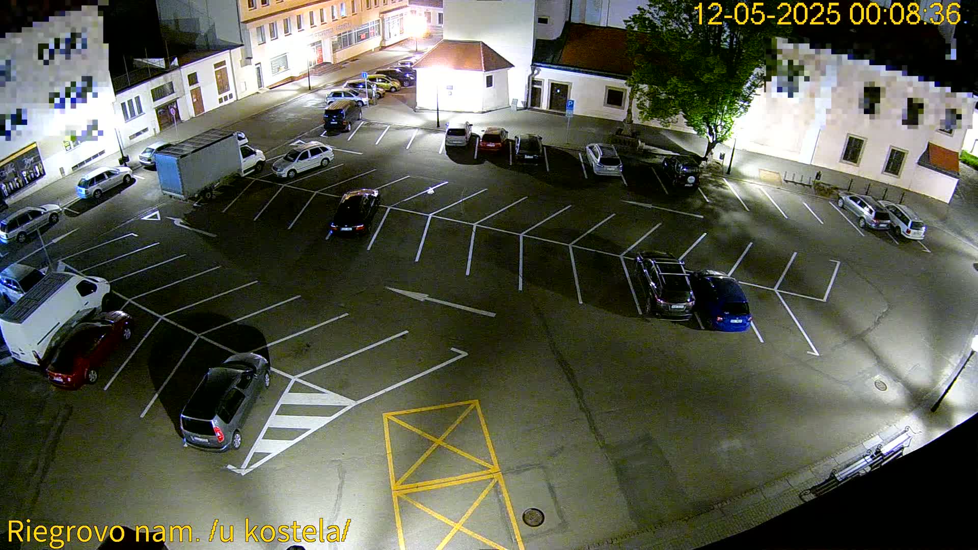 An empty, asphalt parking lot at night, illuminated by artificial lights, contains several parked cars and vans.