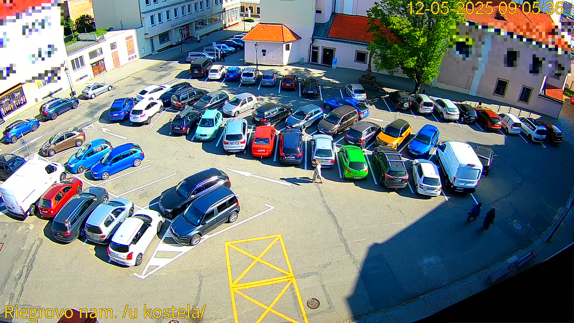 A sunny day reveals a full parking lot with numerous cars parked in various colors, two pedestrians, and a white van.