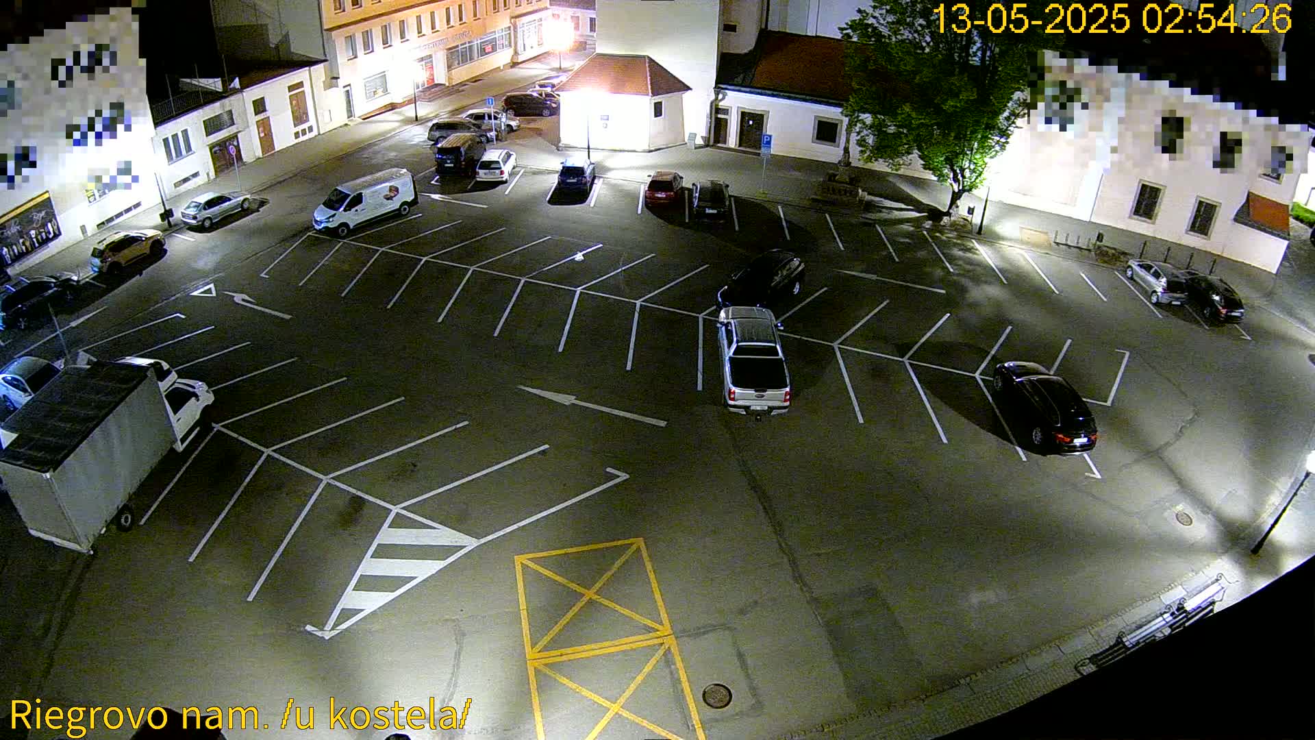 An empty asphalt parking lot at night, with several cars parked within the marked spaces, is illuminated by artificial lights.